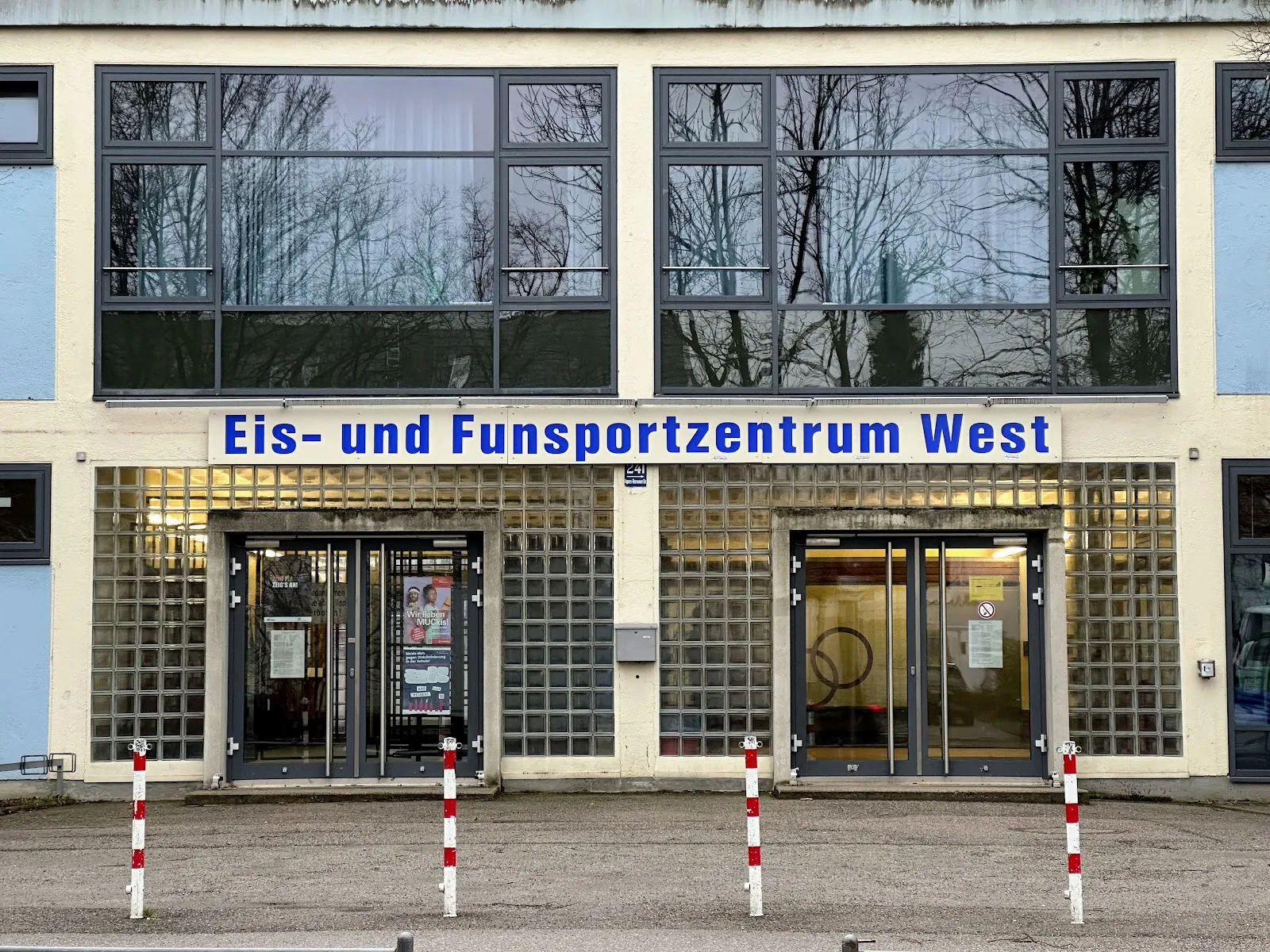 The pale façade with large dark windows and a glass-block wall around the two entrance doors, under the blue Eis- und Funsportzentrum West lettering. — Städtisches Eis- und Funsportzentrum West