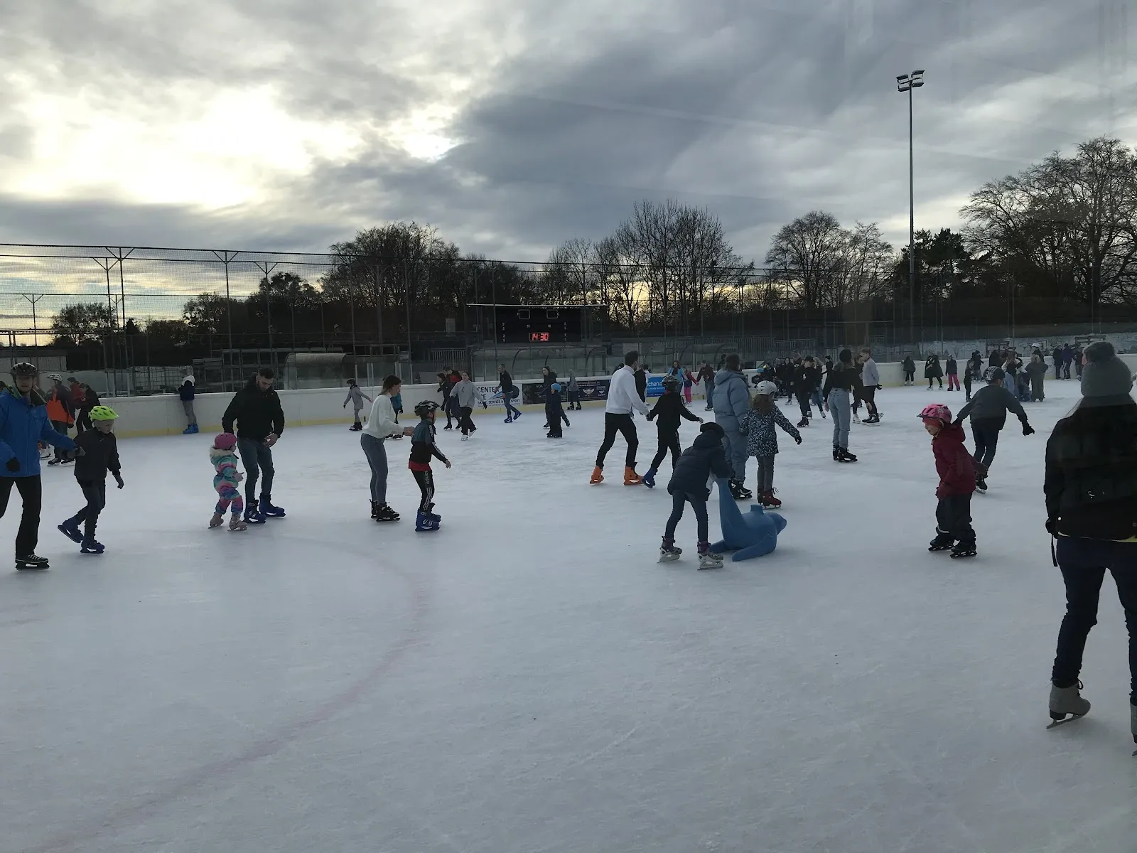 Skaters across the open-air ice of the Eis- und Funsportzentrum West in Munich under a cloudy sky, with the floodlight masts behind. — Städtisches Eis- und Funsportzentrum West