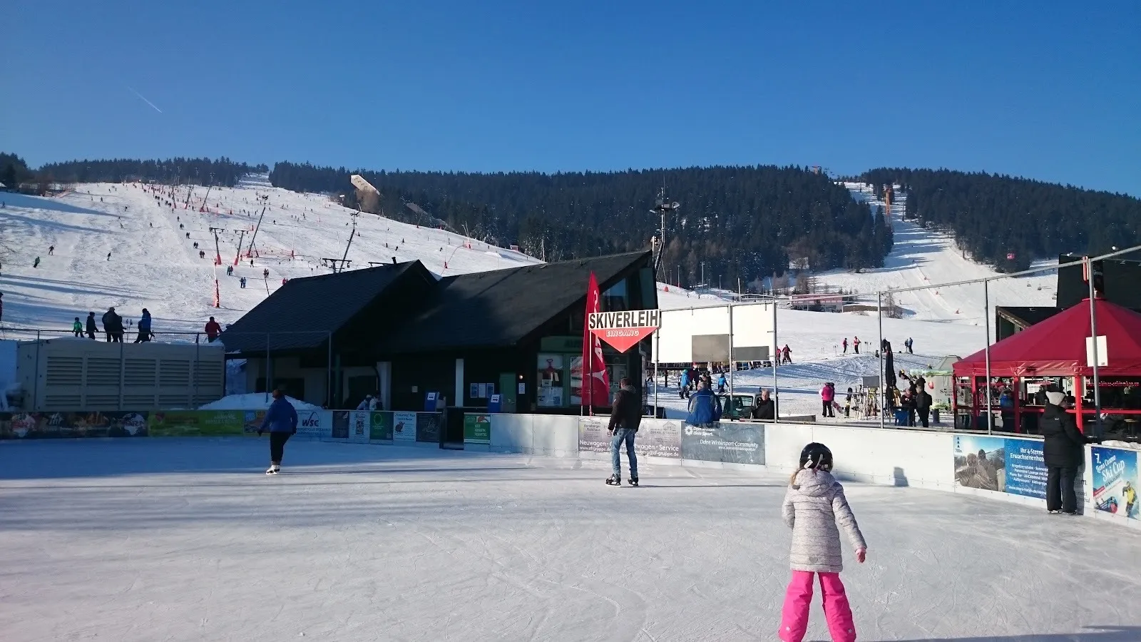 The open-air Oberwiesenthal rink in winter sunshine, skaters on the ice with the clubhouse and the ski slope rising behind. — Eis- und Sportarena Oberwiesenthal