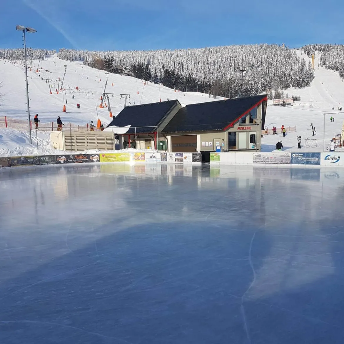 The outdoor ice surface of the Eis- und Sportarena Oberwiesenthal, empty, with the arena building and the ski slope behind it — Eis- und Sportarena Oberwiesenthal