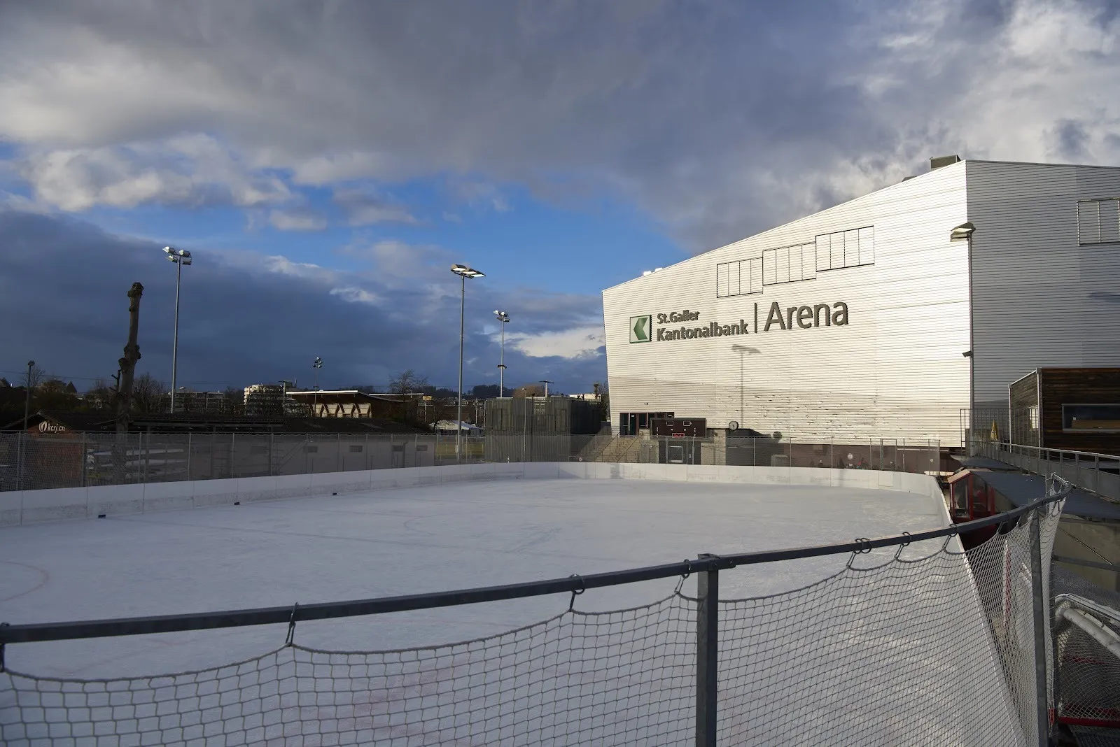 The outdoor Lido ice field beside the white Kantonalbank Arena in Rapperswil, seen from behind the boards at dusk. — Eisanlagen Lido