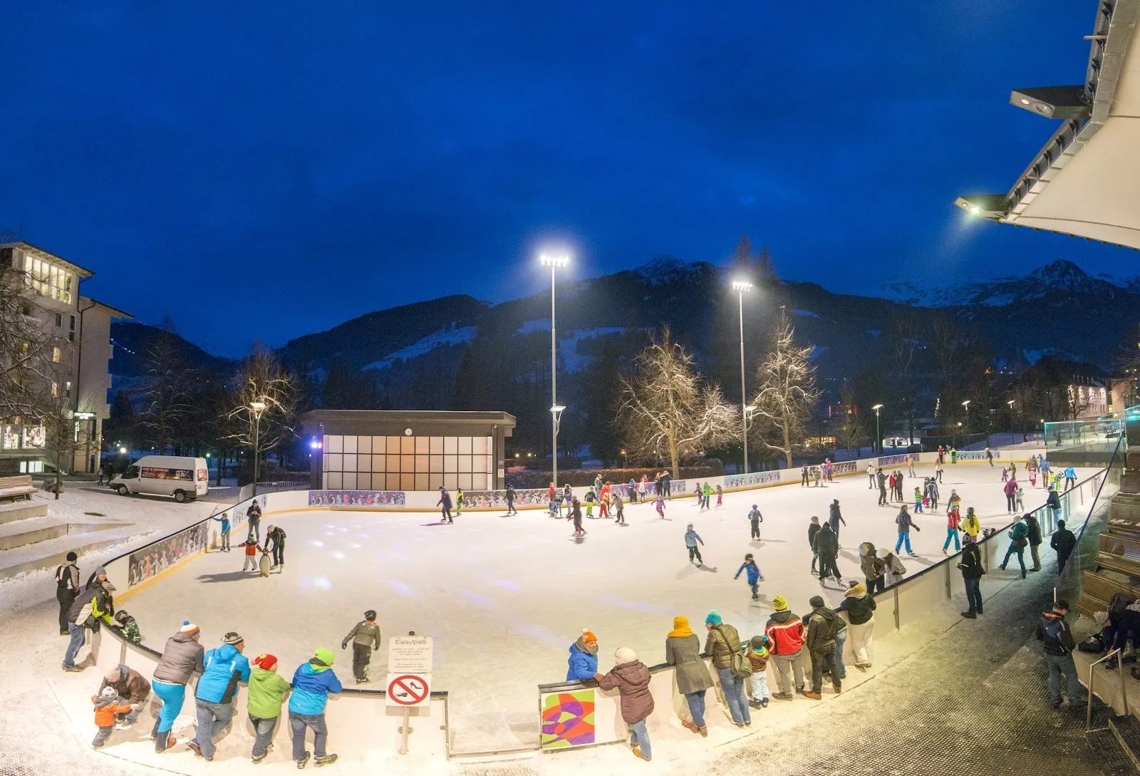Die Freiluft-Eisarena in Bad Hofgastein in der Dämmerung, mit Schlittschuhläufern auf dem Eis unter Flutlicht und den Bergen dahinter. – Eisarena Bad Hofgastein