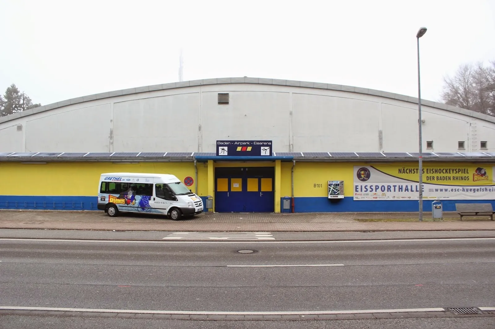 Die Straßenseite der Eisarena am Baden-Airpark: eine lange, flache Halle mit gewölbtem grauem Dach, einer gelb-blauen Front mit den Eingangstüren und dem Schild „Eissporthalle“. – Eisarena am Baden-Airpark