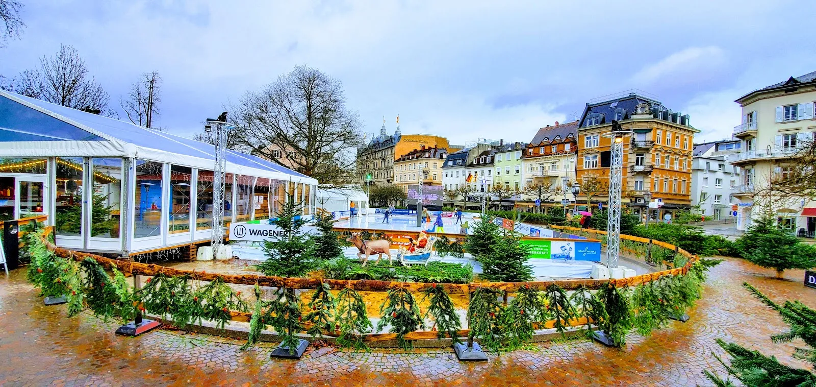 Die saisonale Eisbahn auf dem Augustaplatz, umringt von einem mit Tannengrün geschmückten Zaun, mit einem verglasten Pavillon auf einer Seite und den historischen Gebäuden der Stadt dahinter. – Eisarena Baden-Baden