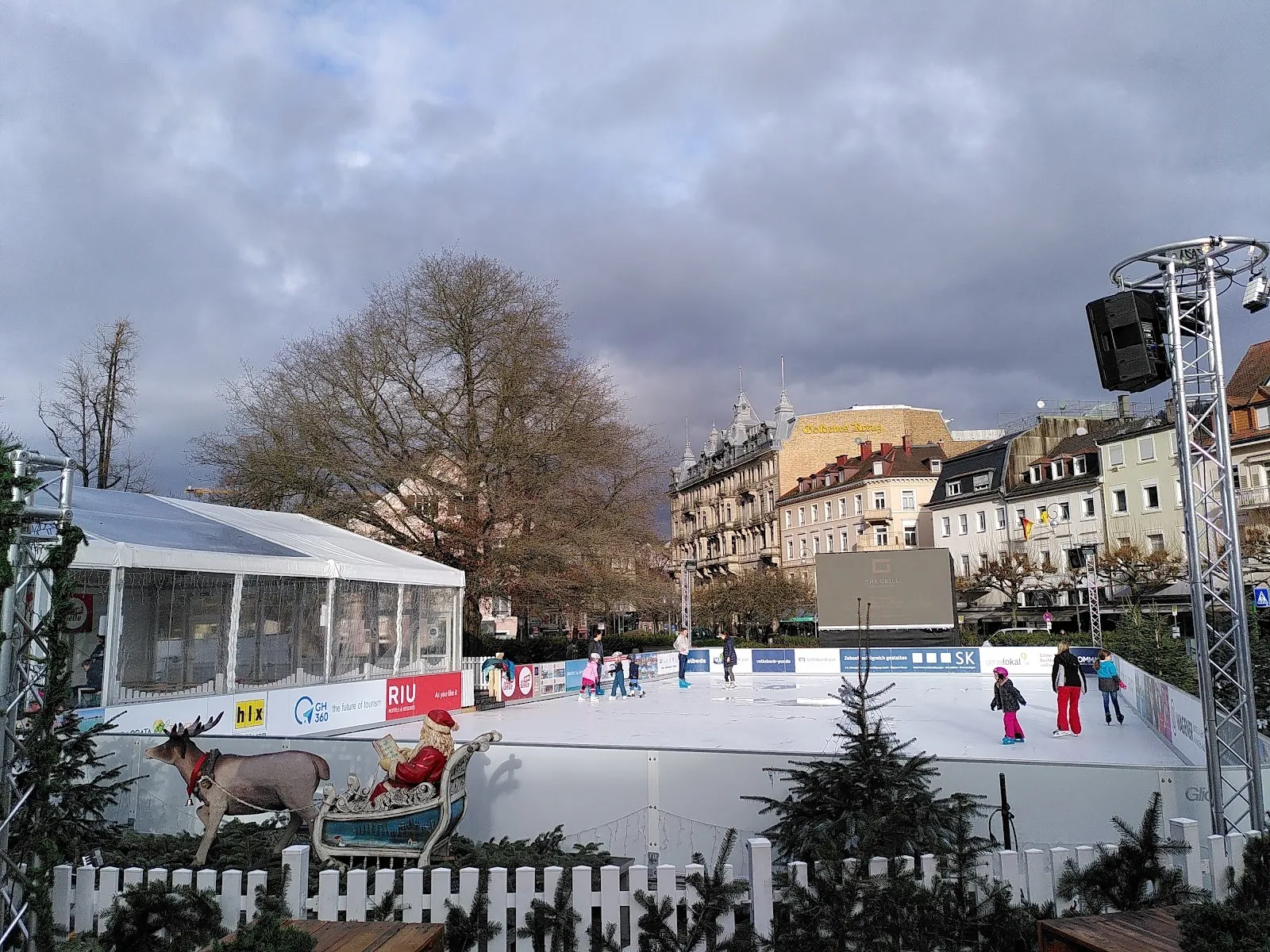 Die saisonale Eisbahn am Augustaplatz in Baden-Baden mit Schlittschuhläufern auf dem Eis und den Gebäuden und Bäumen der Stadt ringsum. – Eisarena Baden-Baden