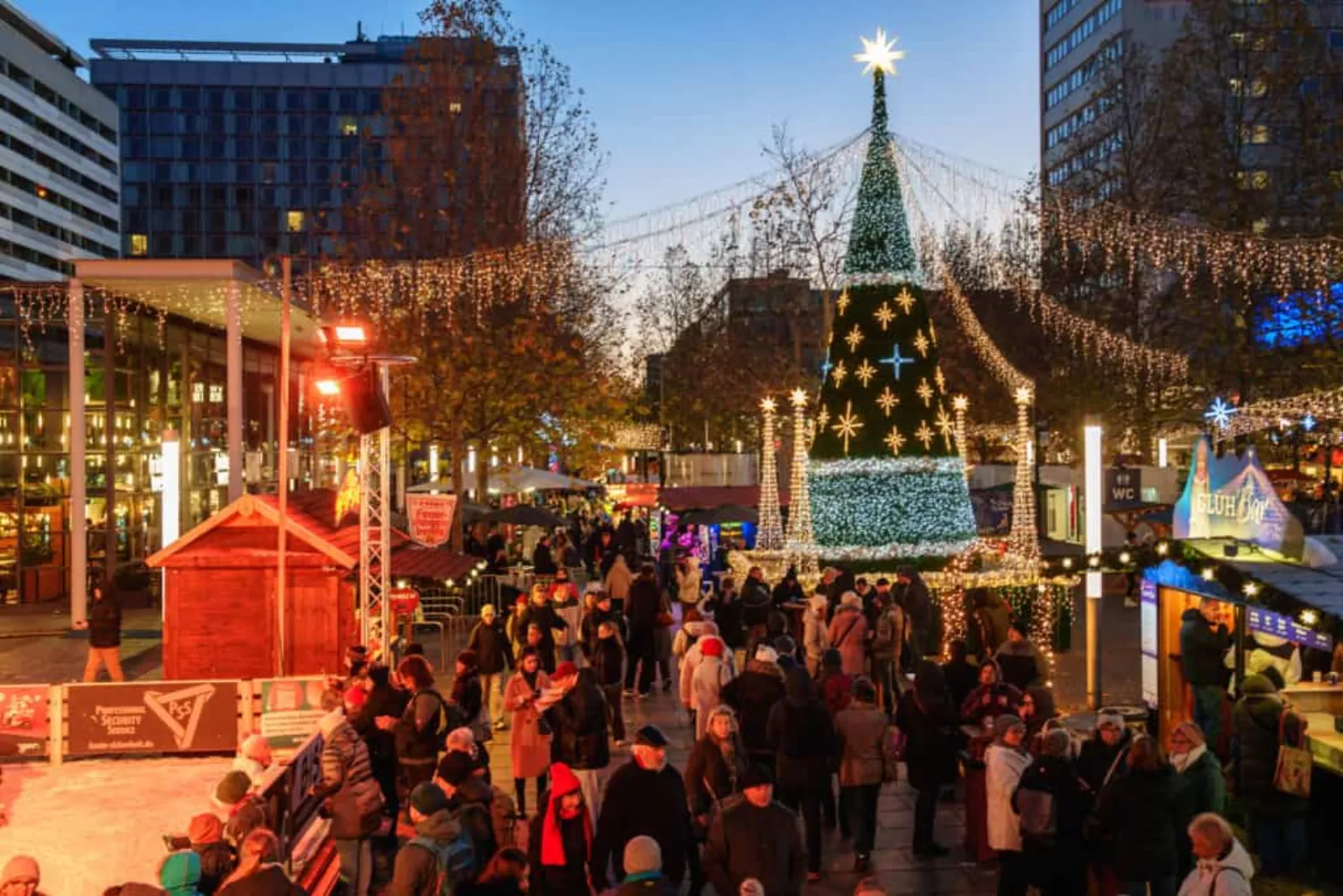 The Dresdner Winterlichter market on Prager Straße at dusk, with the lit Christmas tree, the market stalls and the edge of the rink in the foreground. — Eisarena Dresdner Winterlichter