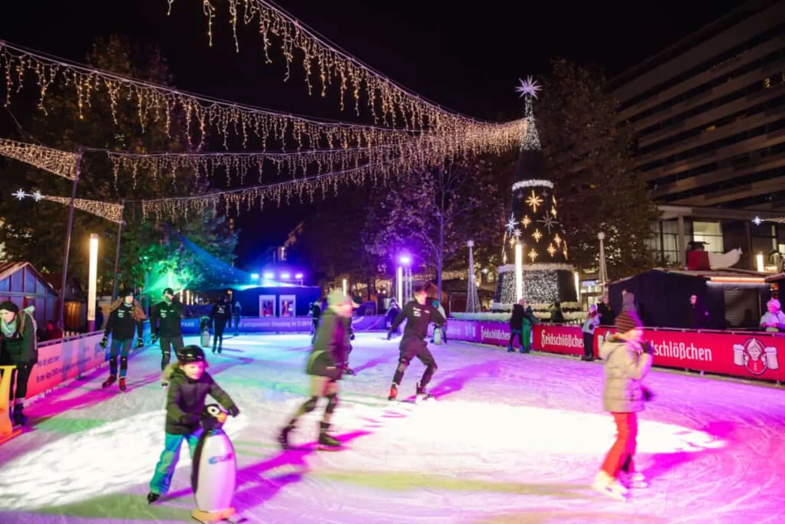 The Eisarena on Prager Straße at night, with skaters of all ages on the lit ice, strings of lights overhead and the advertising boards along the edge. — Eisarena Dresdner Winterlichter
