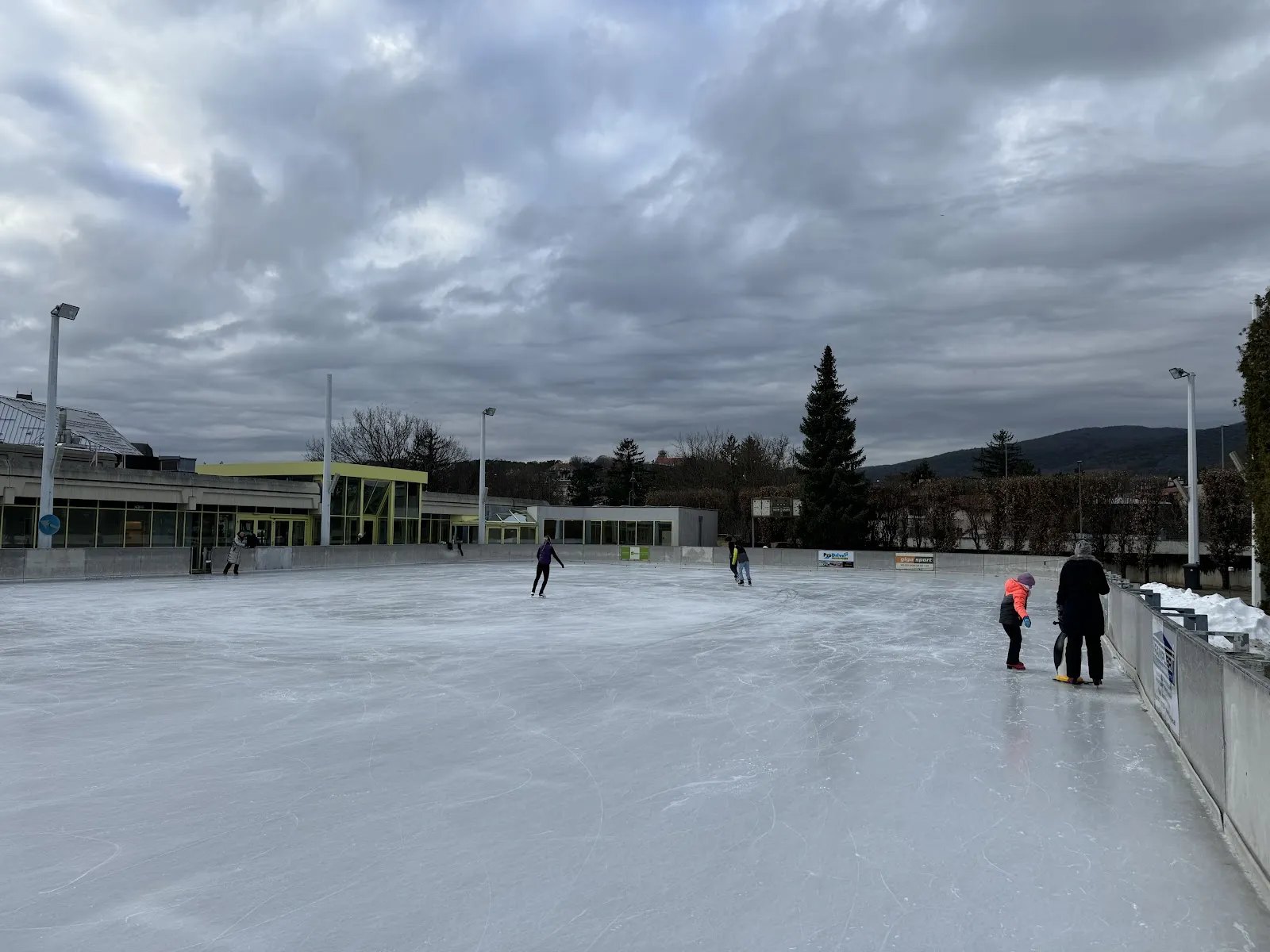 The open-air ice surface at the Freizeitzentrum Perchtoldsdorf under a cloudy sky, with the low pavilion at the edge and a few skaters far across the ice — Eisarena Freizeitzentrum Perchtoldsdorf