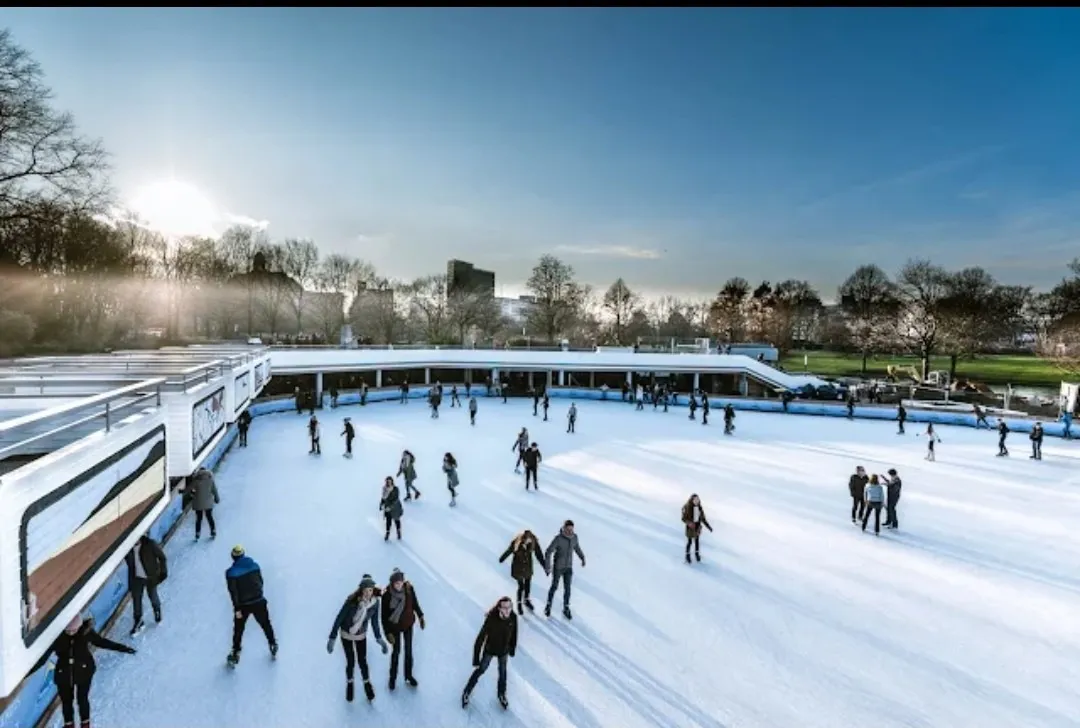 Skaters on the open-air ice of the EisArena Hamburg, seen from above with the park trees and city skyline behind the boards. — EisArena Hamburg