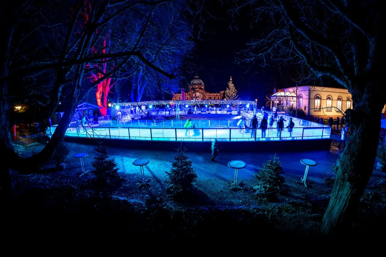 Die Eisbahn im Kurpark von Bad Homburg bei Nacht, blau beleuchtet, mit dem Kurhaus und Bäumen dahinter; Schlittschuhläufer klein in der Ferne. – Eisarena Homburger Weihnachtsdorf