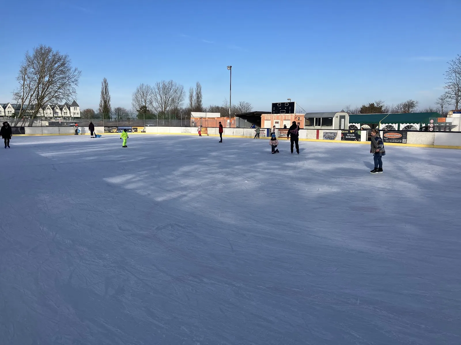The open-air ice surface at Heringsdorf in winter sun, with a few skaters and the low buildings and bare trees of the dune behind the boards. — Eisarena Insel Usedom