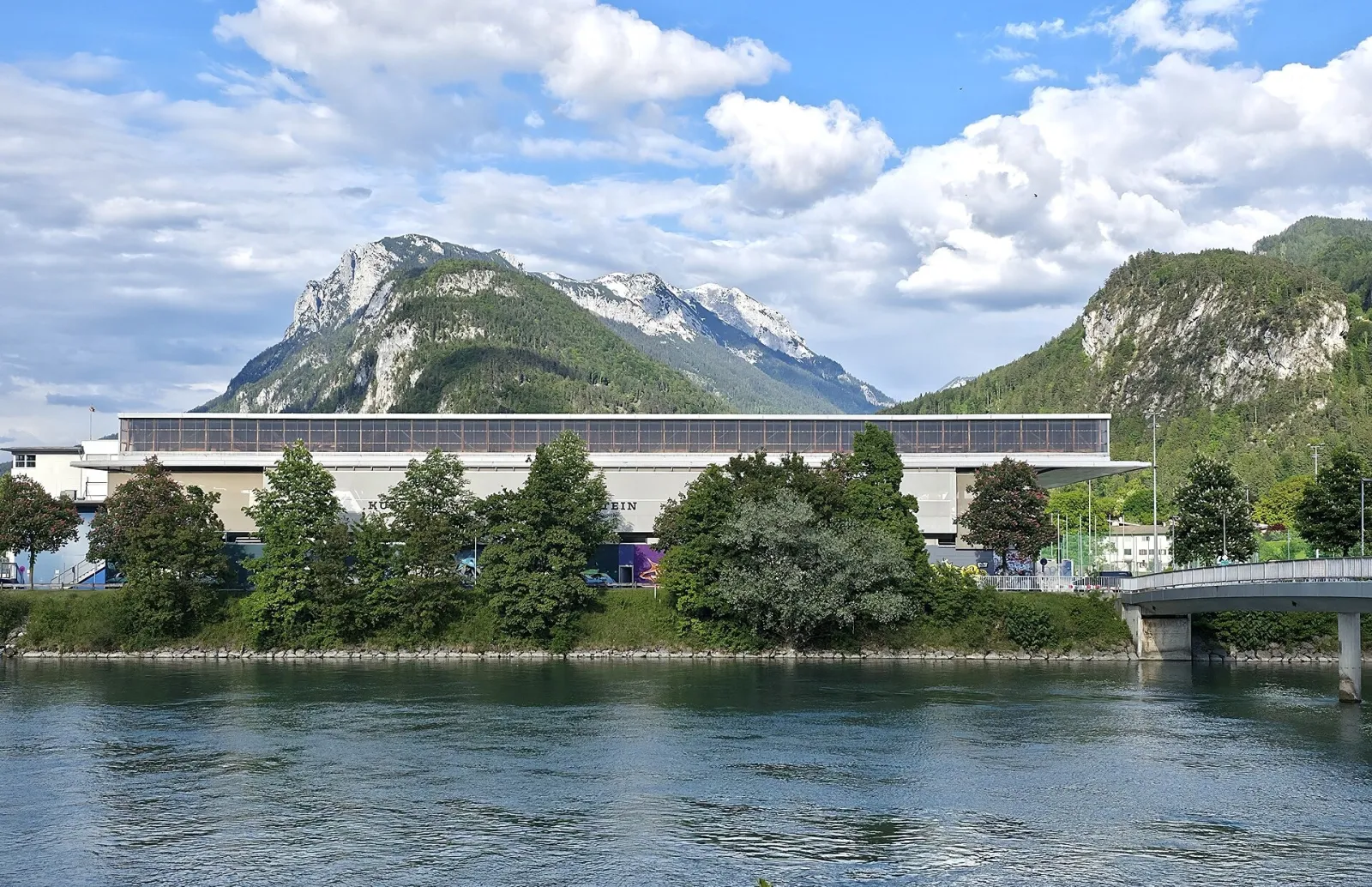 Die Sporthalle Kufstein Arena, über den Inn gesehen, mit den Bergen hinter der langen niedrigen Dachlinie. – Eisarena Kufstein