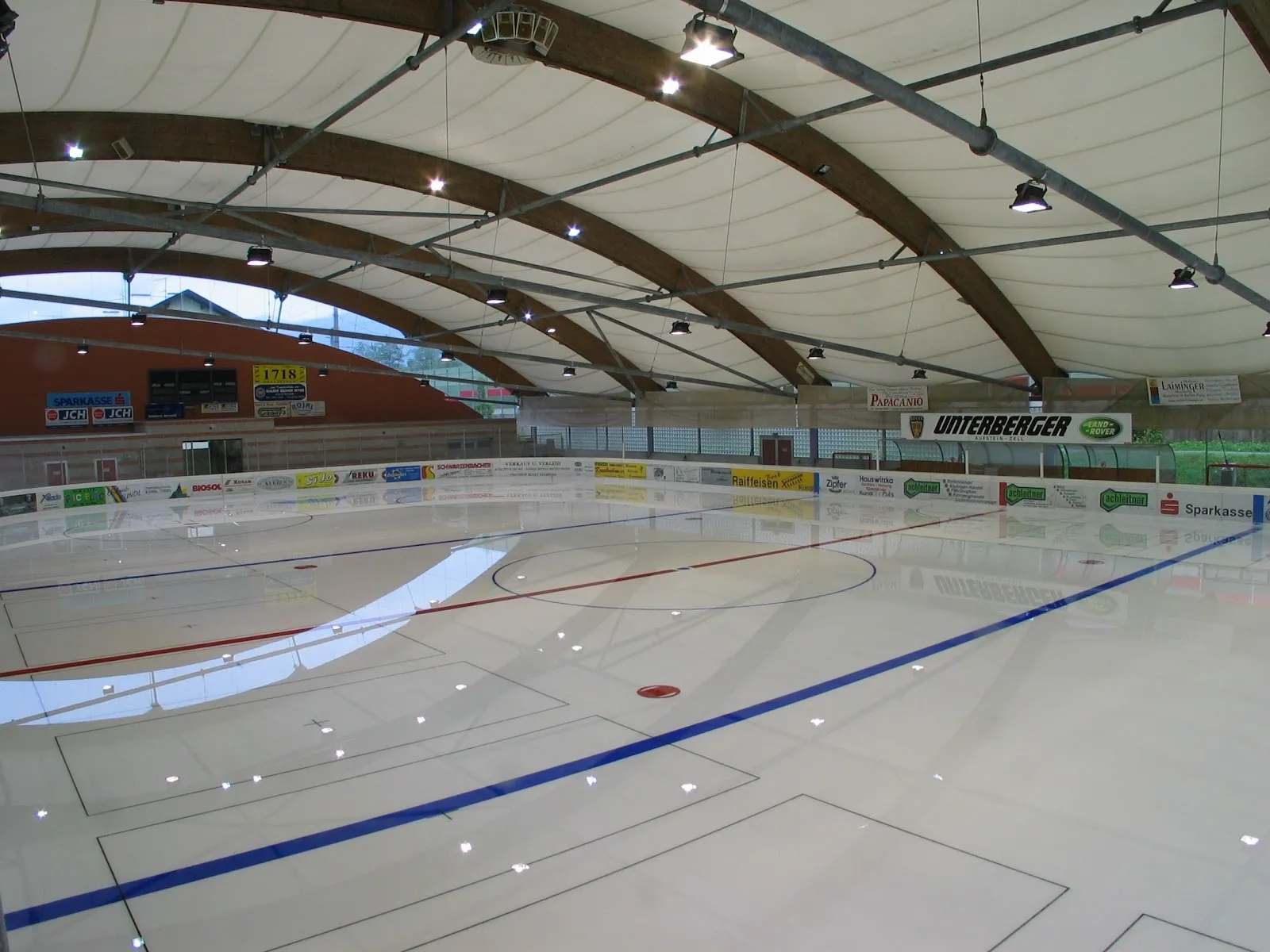 Die Eisfläche der Eisarena Kundl unter ihrem Holzdach mit den blauen Linien und der Bande rund um die Fläche. – Eisarena Kundl