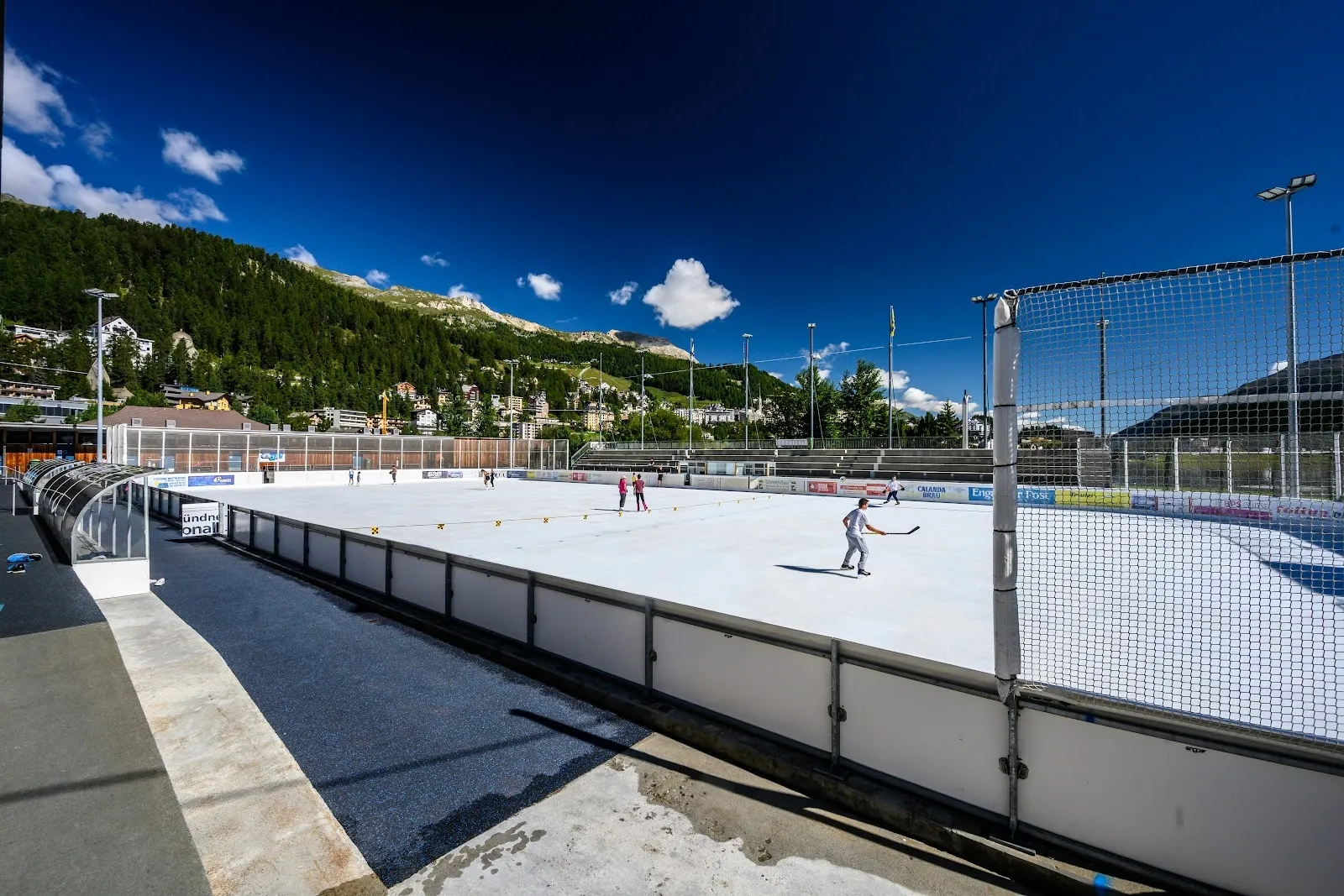 Das Freiluft-Eis der Eisarena Ludains in St. Moritz an einem sonnigen Tag, mit der Bande im Vordergrund und dem bewaldeten Berghang dahinter. – Eisarena Ludains