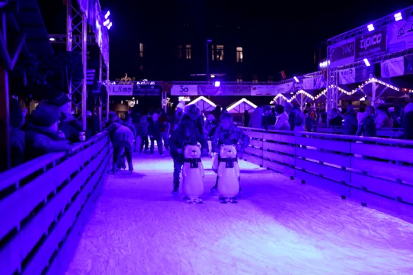 Die Eisarena auf dem Saarlouiser Weihnachtsmarkt am Abend, mit Eisläufern und Pinguin-Laufhilfen auf dem violett beleuchteten Eis hinter einer Holzbande. – Eisarena Weihnachtsmarkt Saarlouis