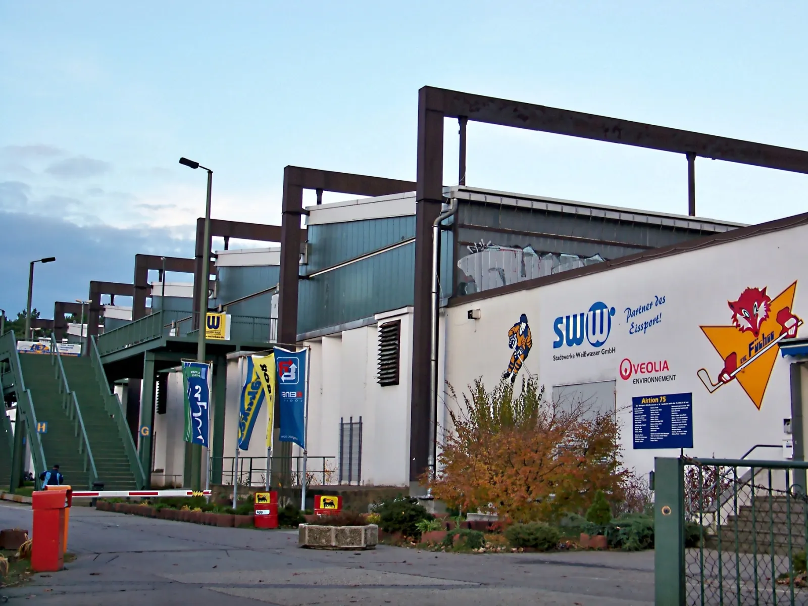 The side of the Weisswasser ice arena with the steel stand frames, Lausitzer Füchse artwork on the wall and sponsor panels by the entrance. — Eisarena Weißwasser