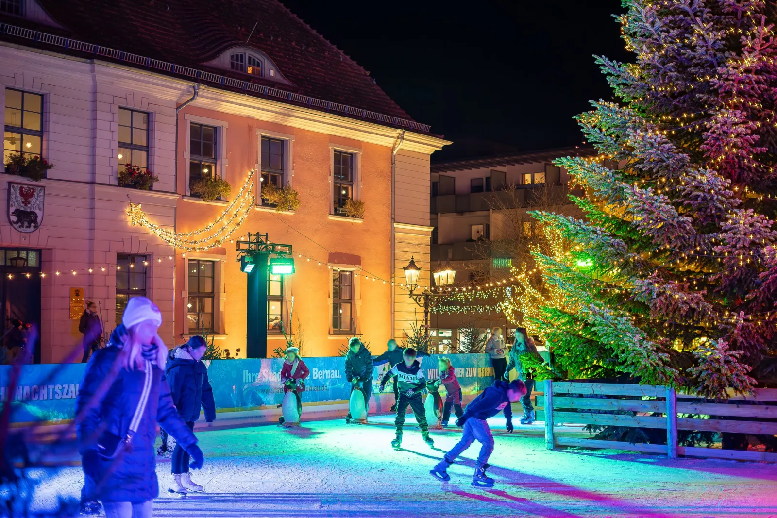 Children and teenagers skating at night on the lit rink on Bernau's Marktplatz, with penguin skating aids, the decorated tree and the old town hall behind. — Eisbahn zum Bernauer Weihnachtsmarkt