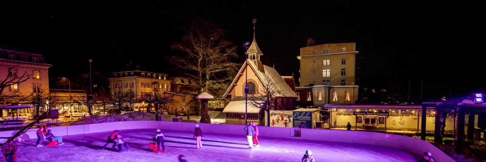 The lit ice field on the Casinoplatz at night, with a few skaters and the village chapel and houses behind it — Kunsteisbahn Meiringen (Casinoplatz)