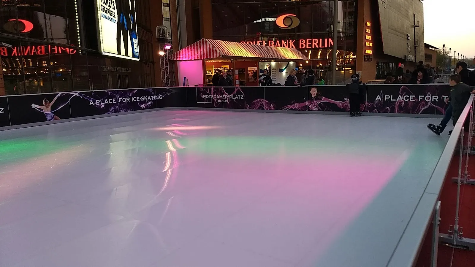 The small synthetic ice rink at dusk, lit in pink and green, with decorated boards, a wooden hut and the Potsdamer Platz buildings glowing behind. — Eisbahn im Center am Potsdamer Platz
