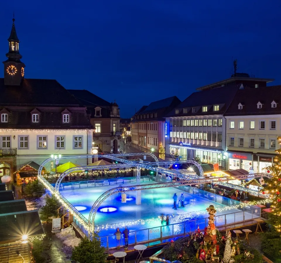 Die Eisbahn auf dem Marktplatz in Emmendingen in der Dämmerung, mit der beleuchteten Eisfläche vor den historischen Gebäuden und einem Kirchturm. – Eisbahn Emmendingen