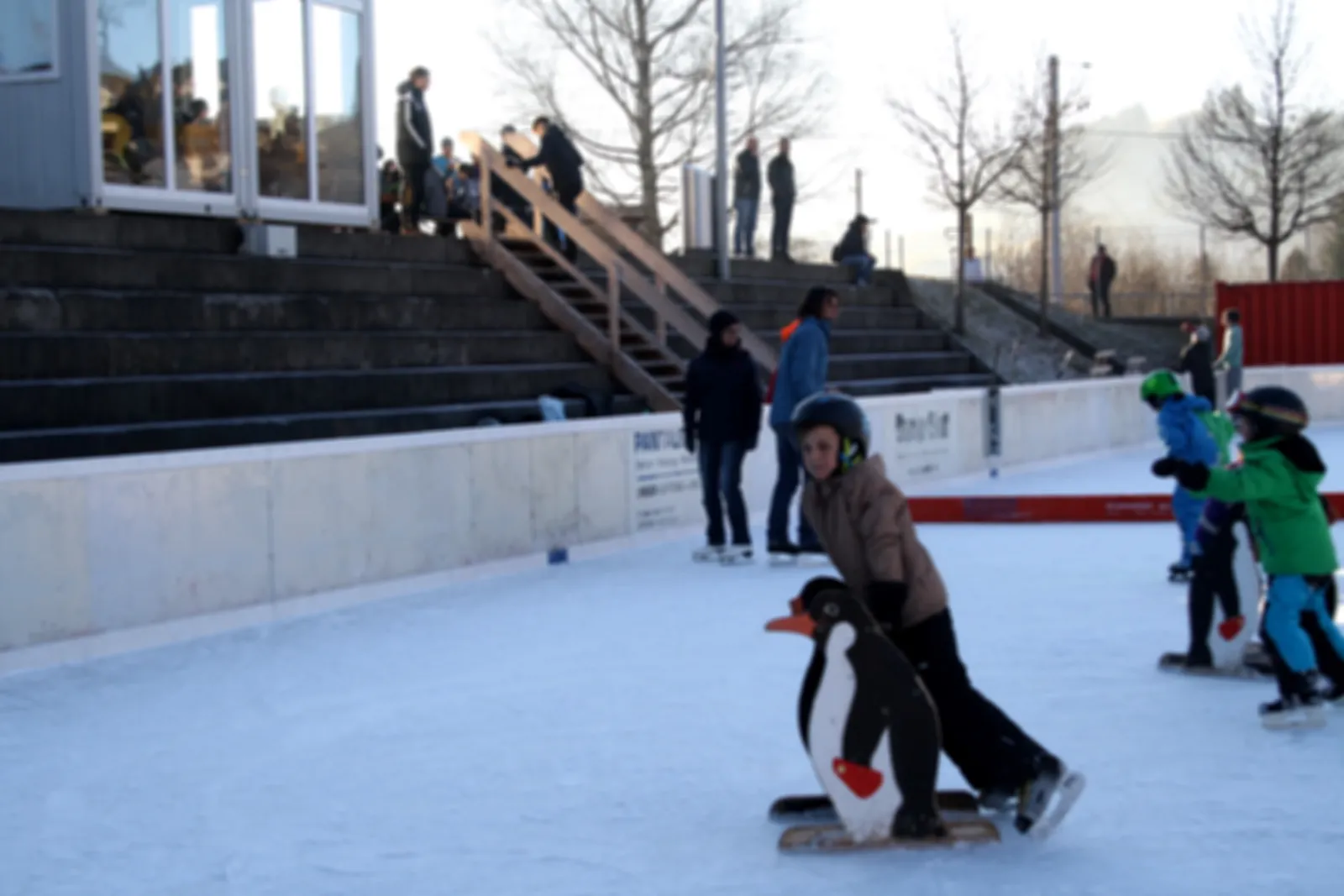 Children skating on the Eisbahn Gais ice beside the steps up to the rink, with a helper penguin figure and spectators at the top. — Eisbahn Gais
