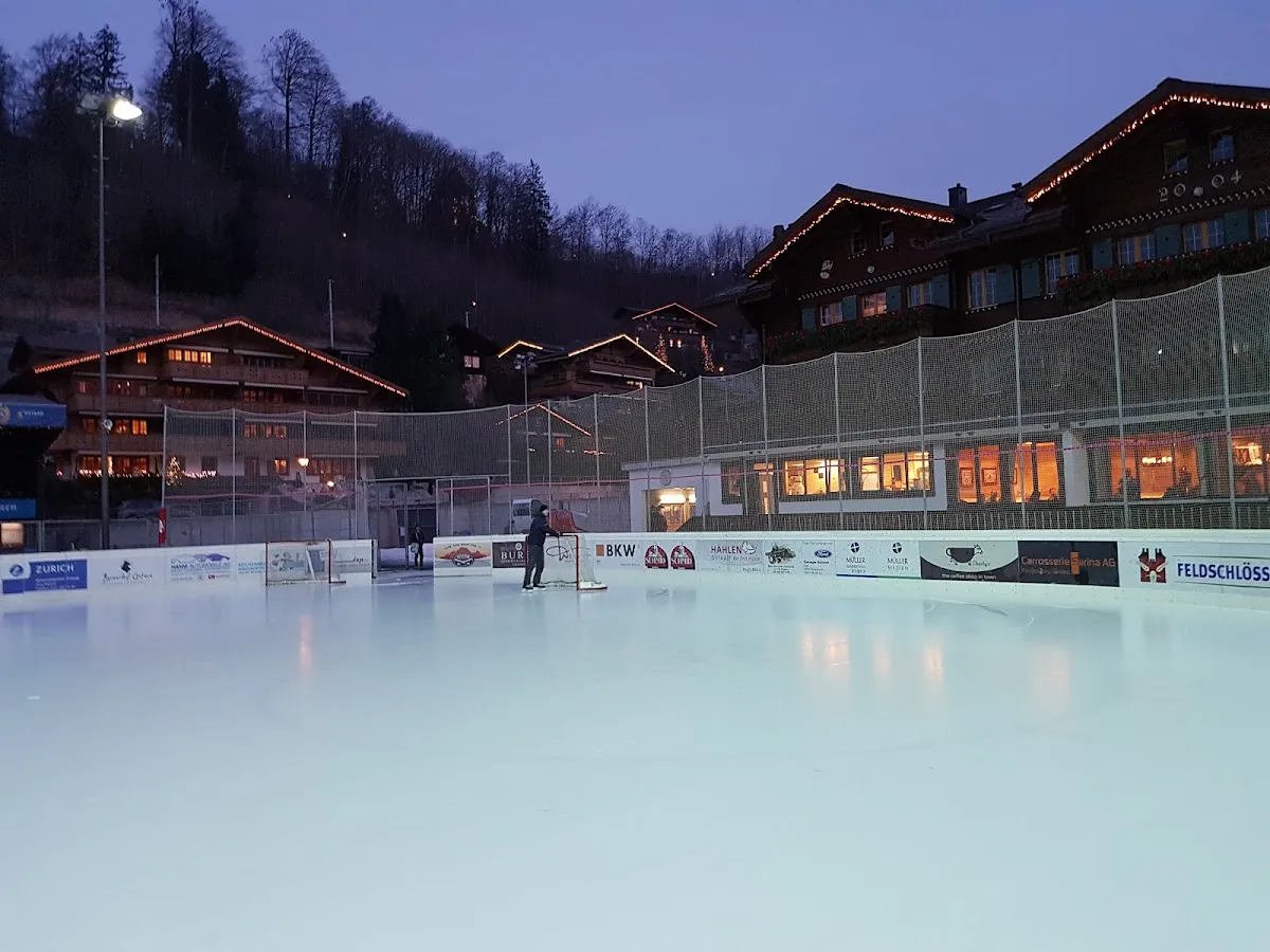 Das Eisfeld der Eisbahn Gstaad in der Dämmerung, mit den Banden und den Chalets des Dorfs dahinter – Eisbahn Gstaad