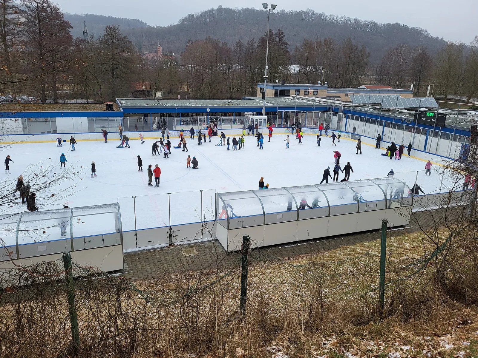 Die Freiluft-Eisfläche von der Tribünenseite aus gesehen, mit überdachten Bänken und einigen kleinen Schlittschuhläufern in mittlerer Entfernung. – Kunsteisbahn Kulmbach