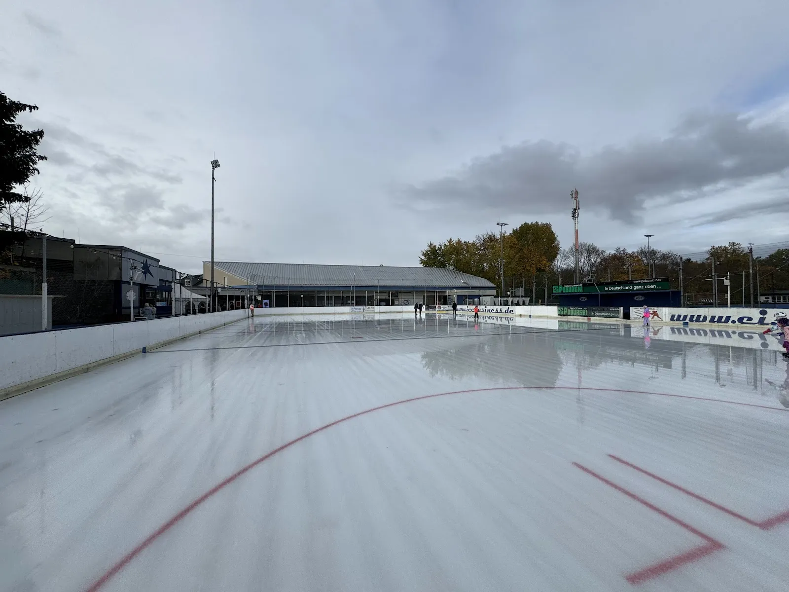 Die Freiluft-Eisfläche erstreckt sich unter bewölktem Himmel, mit roten Linienmarkierungen, niedrigen Banden und dem langen überdachten Gebäude dahinter. – Eisbahn Lankwitz