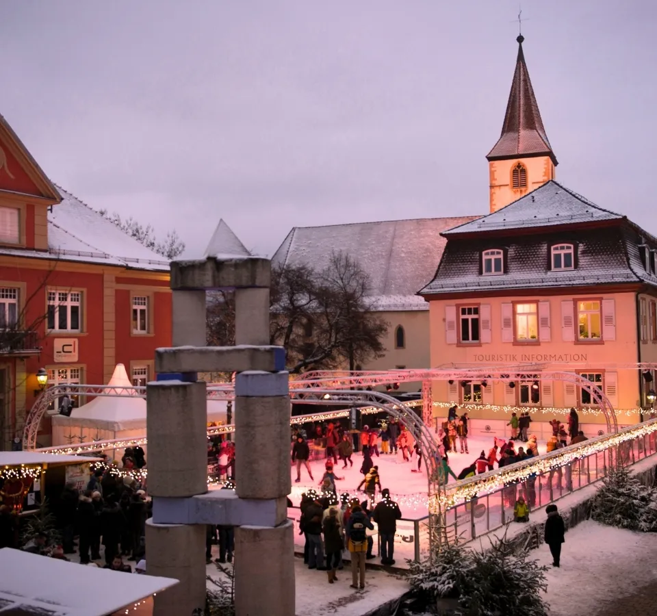 The ice rink on the Markgräfler Platz in Müllheim at dusk, with skaters on the lit ice in front of the church and the houses around the square. — Eisbahn Müllheim