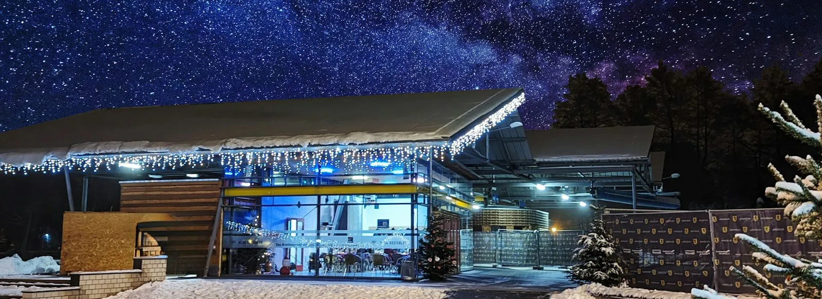 The glass-fronted IGLU pavilion beside the Eisbahn at night under a starry sky, with snow and Christmas trees around it. — Eisbahn Oelsnitz/V.
