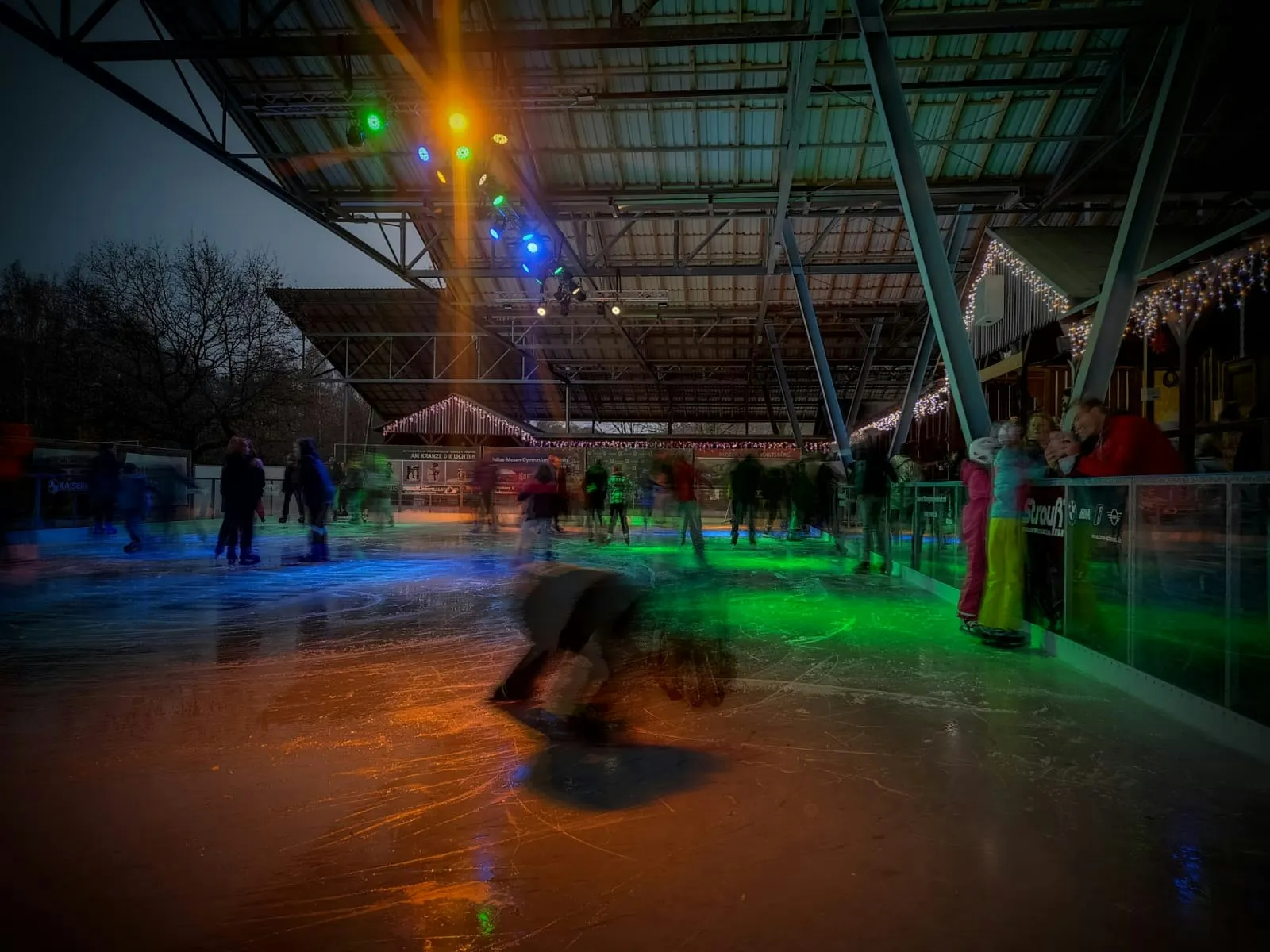 Skaters on the lit ice under the open roof of the Eisbahn at night, with the log-cabin stalls behind. — Eisbahn Oelsnitz/V.