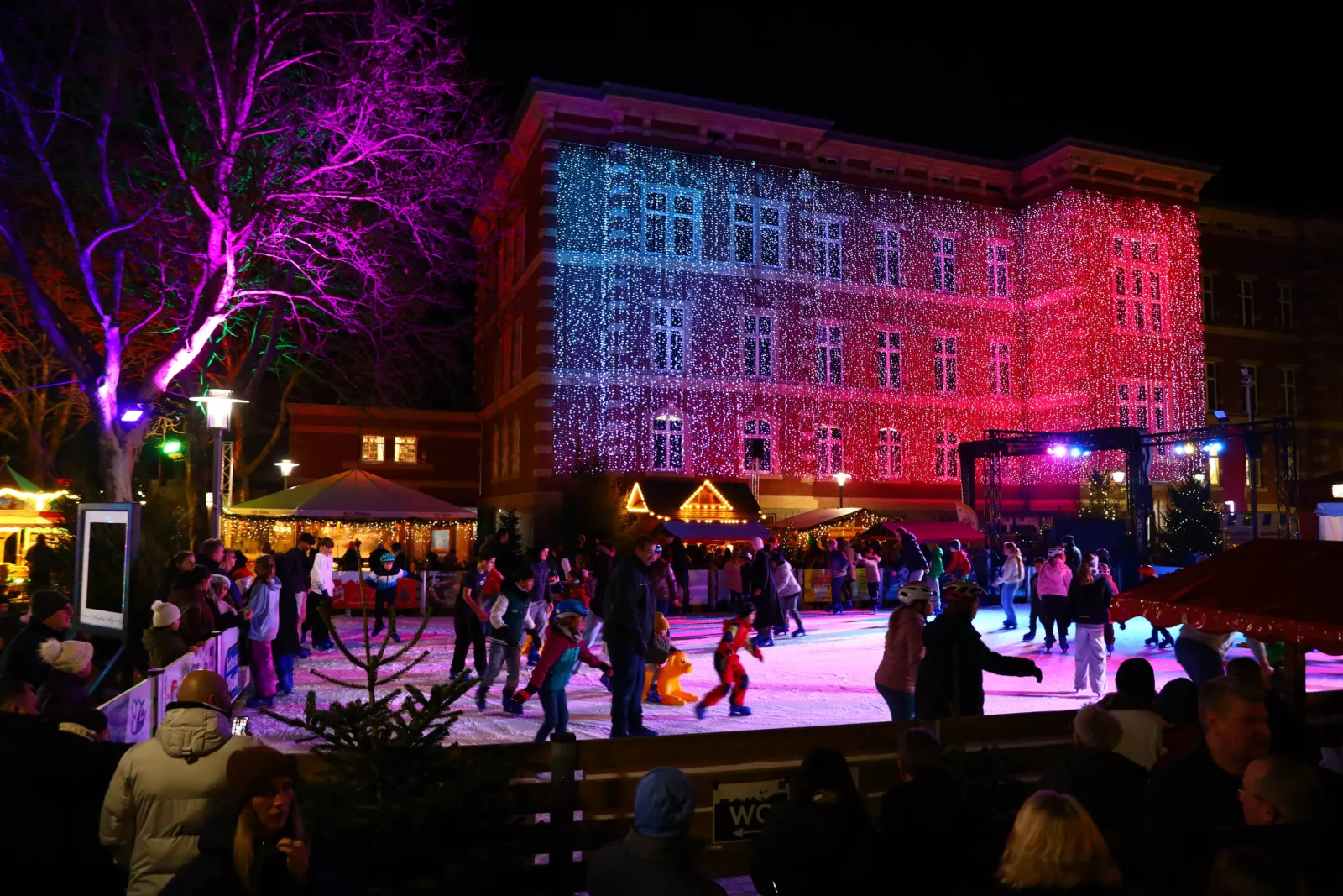 Schlittschuhläufer auf der Eisbahn des Seesener Lichterzaubers bei Nacht, vor dem rot und blau beleuchteten Jacobson-Haus und umgeben von Marktständen. – Eisbahn Seesener Lichterzauber