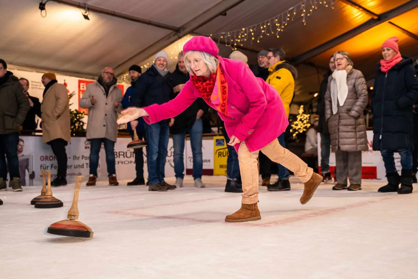 Spielerinnen und Spieler werfen Eisstöcke auf dem Eis unter dem überdachten Zelt, dahinter Zuschauer und Weihnachtsbeleuchtung. – Eisbahn Versmold