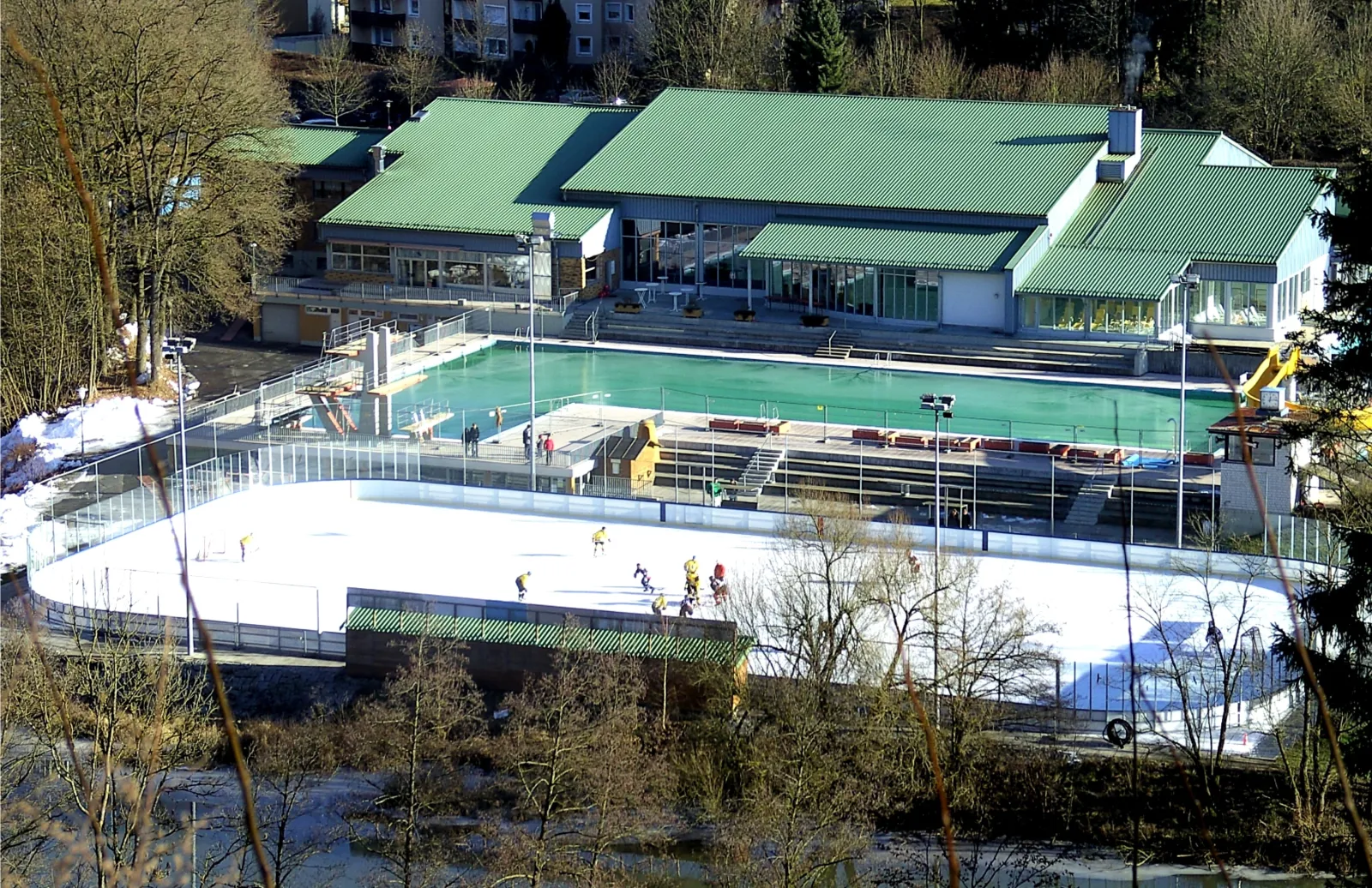 The outdoor ice rink beside the green-roofed Vilshofen pool building, seen from the slope above, with a few skaters on the white ice and bare trees around. — Eisbahn Vilshofen