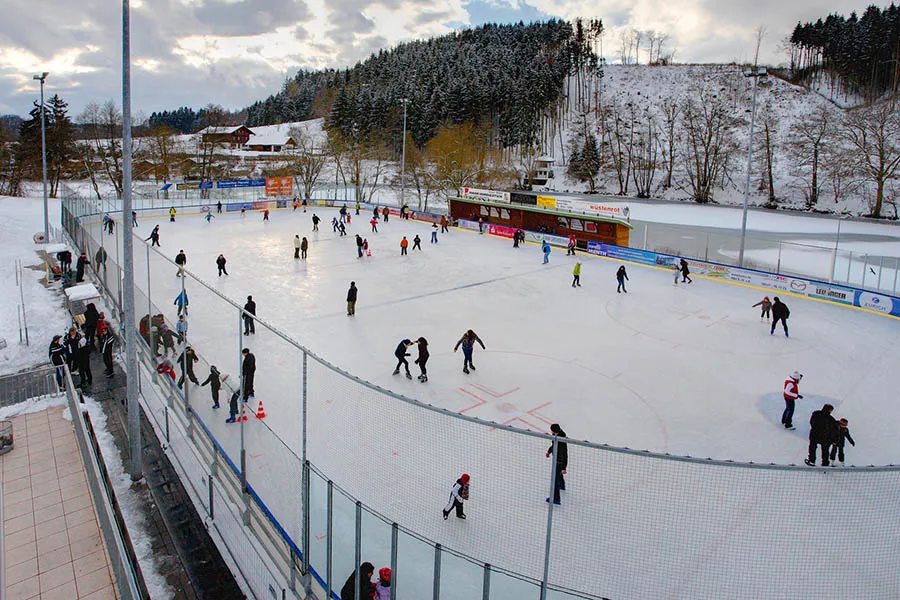 The open-air rink in a snowy valley, with skaters spread across the ice, the boards and the wooden kiosk on the far side. — Eisbahn Vilshofen