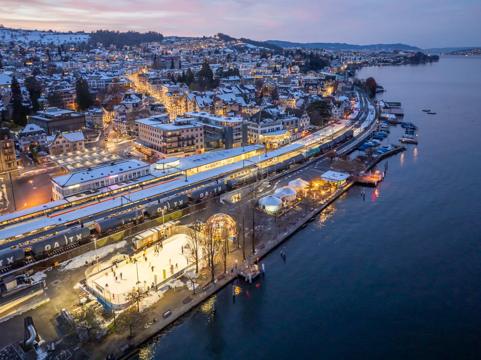 The lit Eisbahn Wädi on the Seeplatz seen from the air at dusk, between the railway station and Lake Zürich, with the old town of Wädenswil behind — Eisbahn Wädi