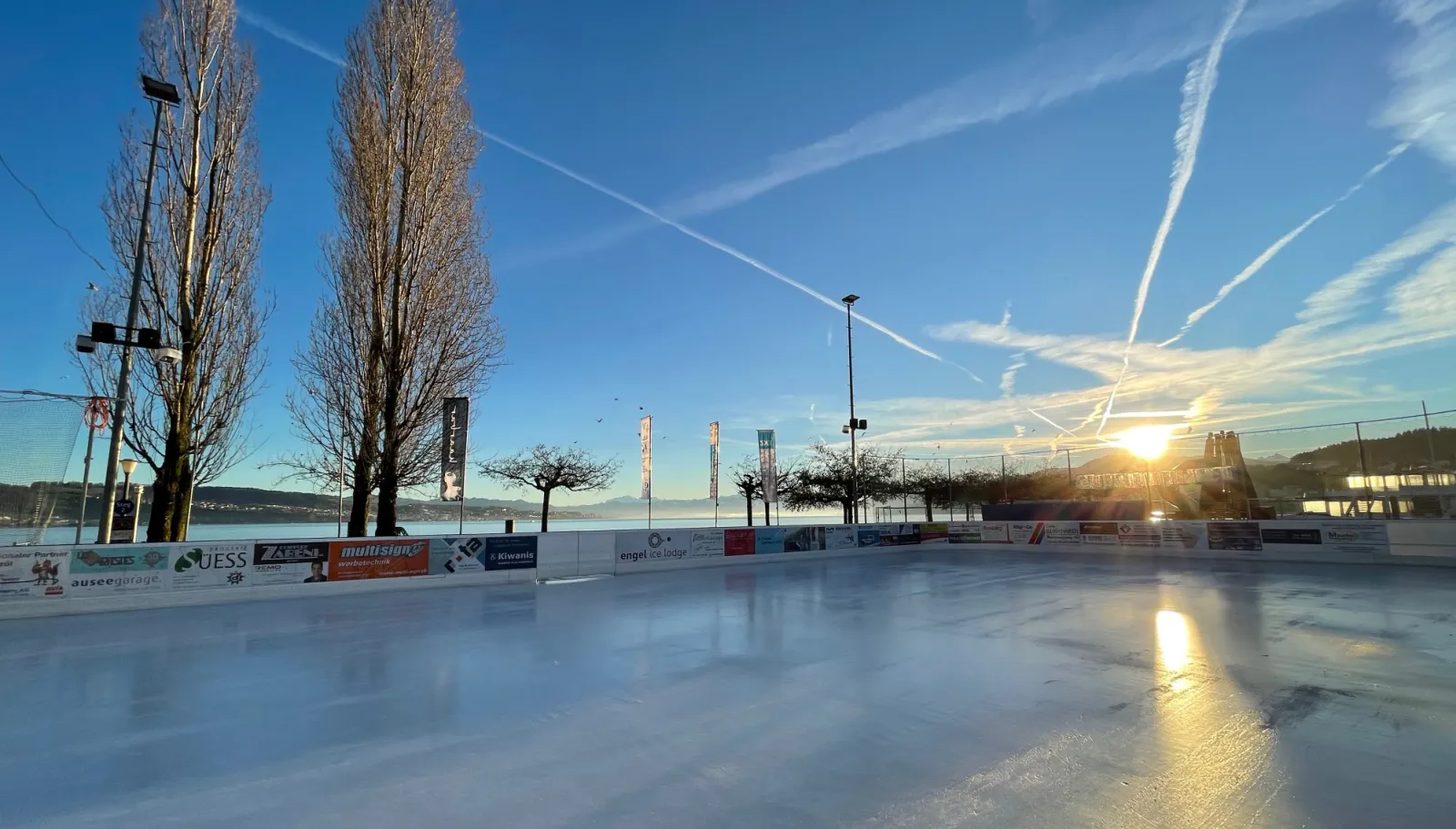 The empty ice field of the Eisbahn Wädi at sunrise, with the lake and the bare trees of the Seeplatz behind the boards — Eisbahn Wädi