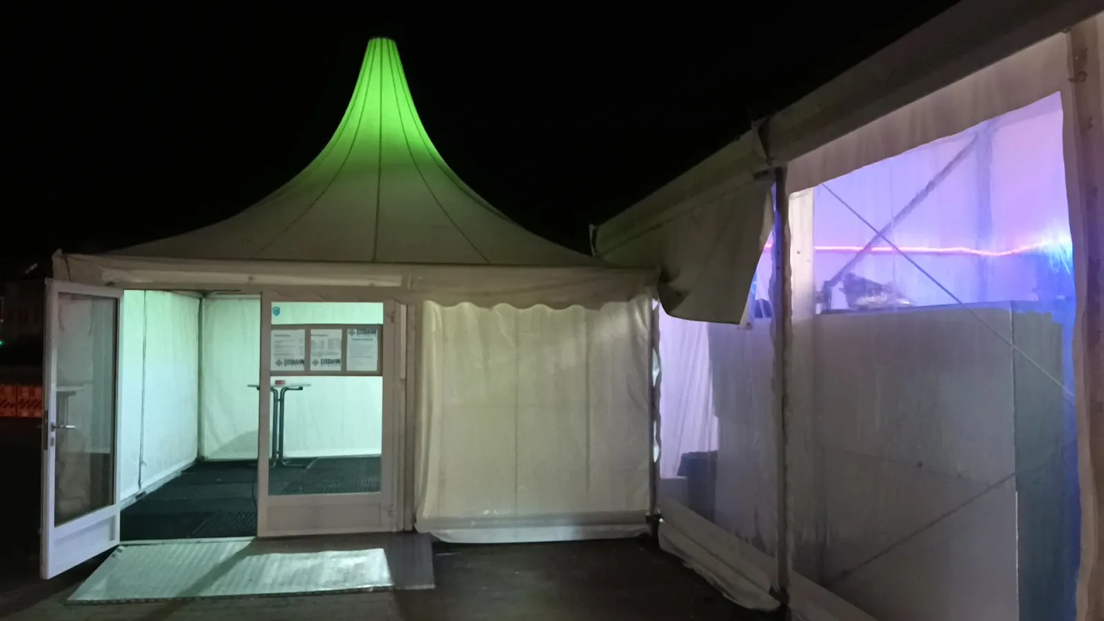Der beleuchtete Zelteingang der Wismarer Eisbahn bei Nacht, mit dem grün leuchtenden Dachfirst des Pavillons neben dem überdachten Eis. – Eisbahn Wismar