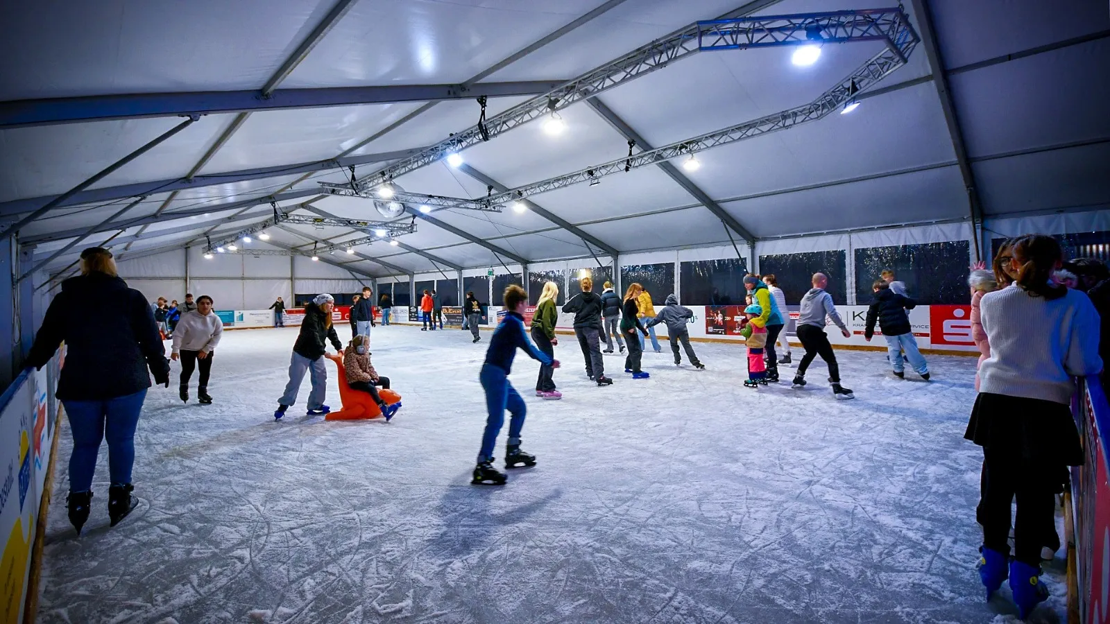 Schlittschuhläufer jeden Alters auf dem beleuchteten Eis unter dem weißen Dach der Wismarer Eisbahn, im Vordergrund ein Kind mit einer orangefarbenen Lauflernhilfe. – Eisbahn Wismar