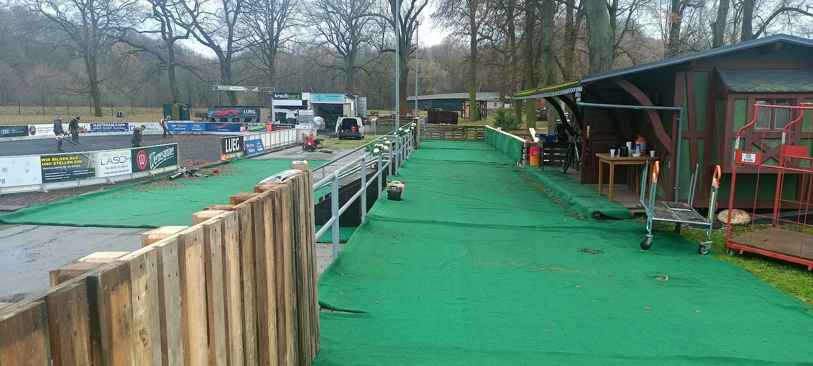 Ein grün ausgelegter Gehweg zwischen den Eisbahnbanden und einer Holzhütte im Park, dahinter die Werbebanden der Bahn und kahle Bäume. – Eisbahn Zwickau im Muldepark