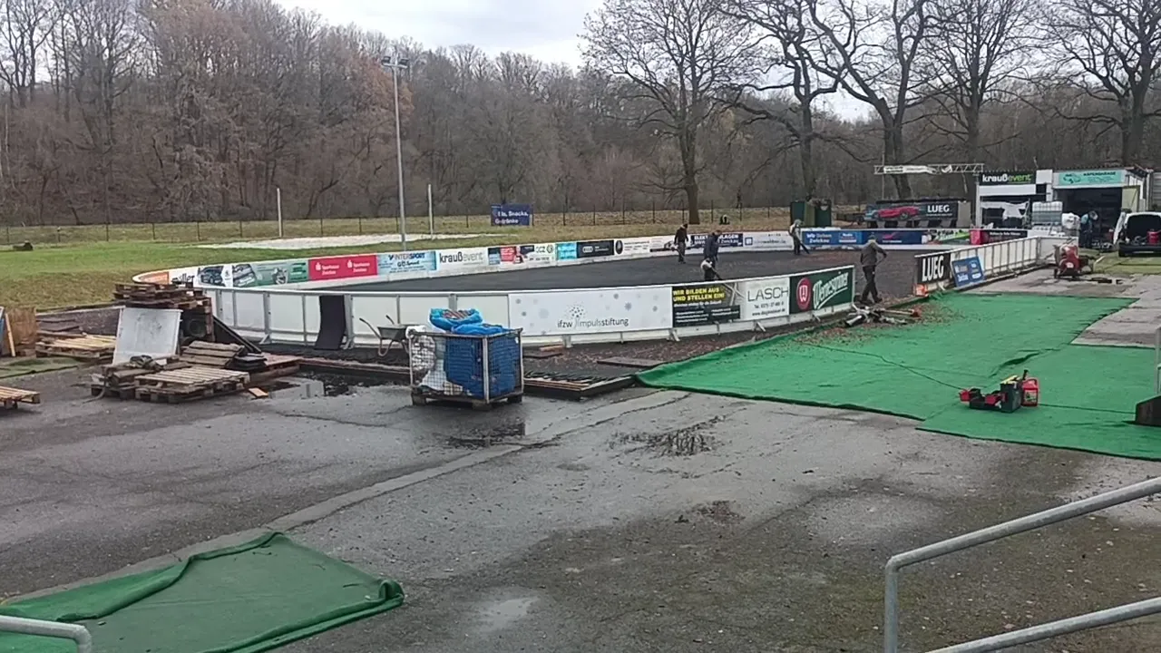 Die Freiluft-Eisbahn im Muldepark bei Tag mit Werbebanden und einem grünen Teppichweg, dahinter kahle Bäume und ein Hang. – Eisbahn Zwickau im Muldepark