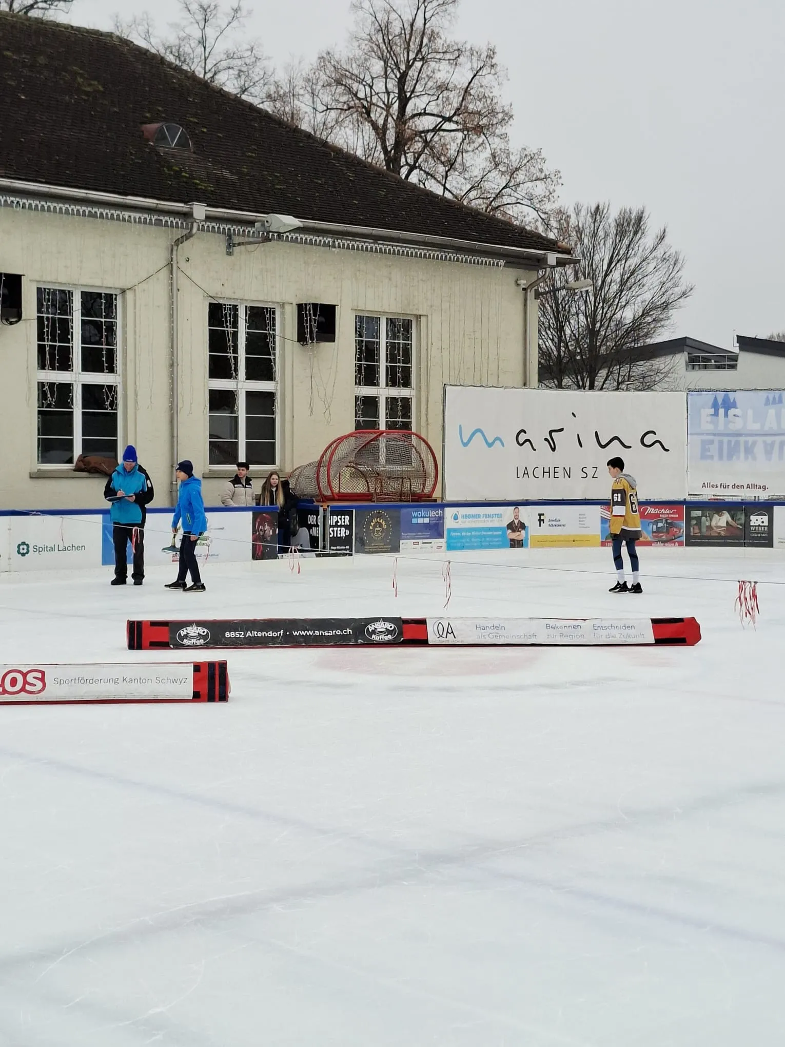 Das Freiluft-Eisfeld Lachen mit dem Marina-Gebäude und Sponsorenbanden; einige Personen auf dem Eis, niemand posiert. – Eisfeld Lachen