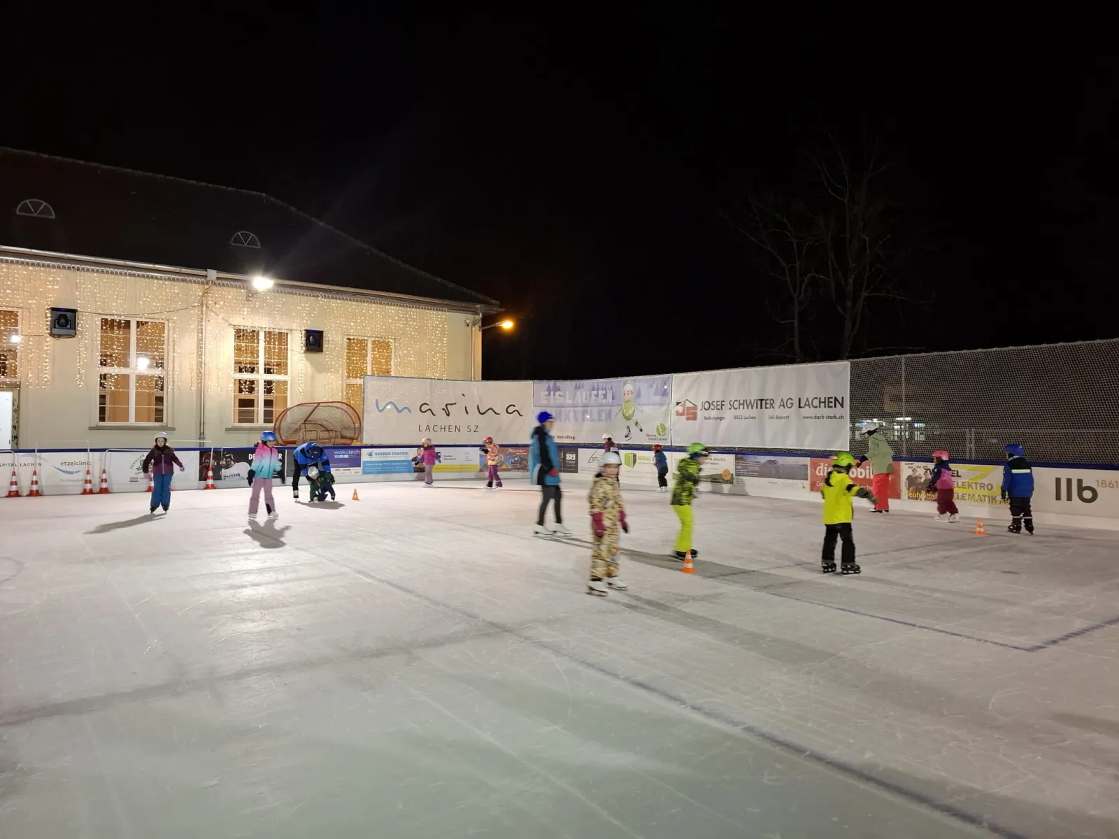 Eisläufer auf dem nachts beleuchteten Eisfeld Lachen, dahinter das Gebäude am See; Eisläufer in der Ferne. – Eisfeld Lachen