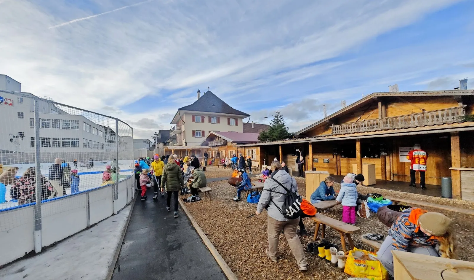 Schlittschuhläufer auf dem Freiluft-Eis des Eisfelds Langnau im Emmental, mit dem Netz, den Gebäuden am Rand und dem Chalet dahinter. – Eisfeld Langnau