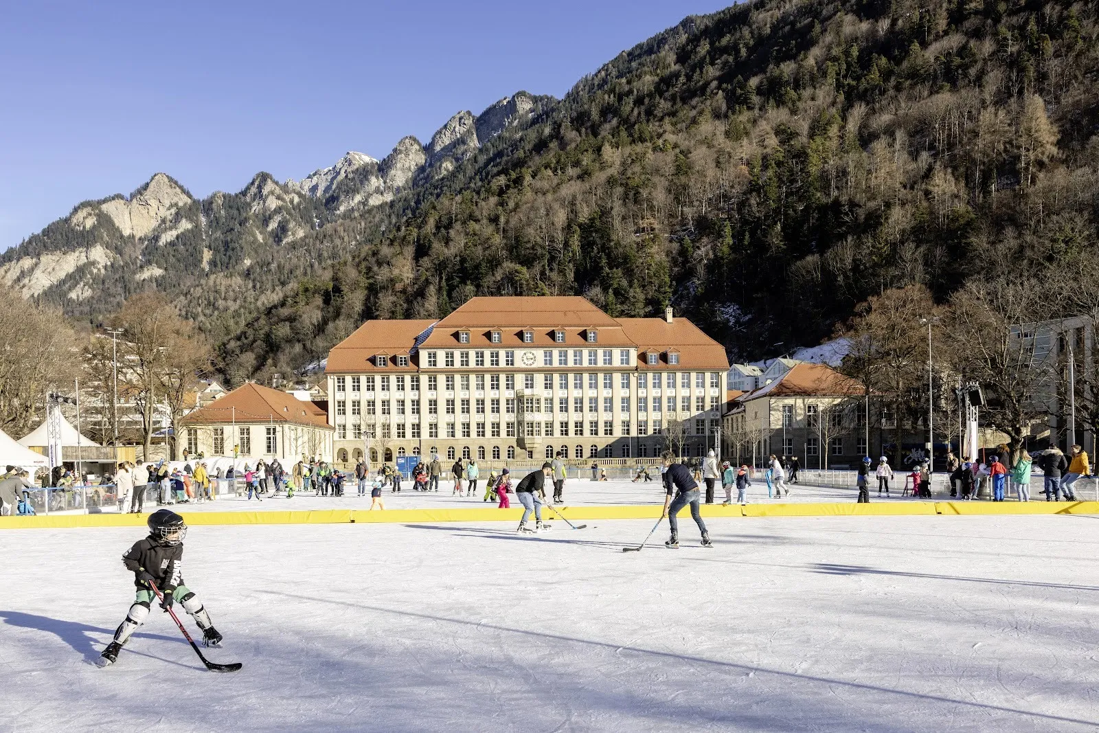 Schlittschuhläufer und ein junger Spieler auf dem Freiluft-Eisfeld Quader in Chur, dahinter das lange Schulgebäude und die Berge. – Eisfeld Quader