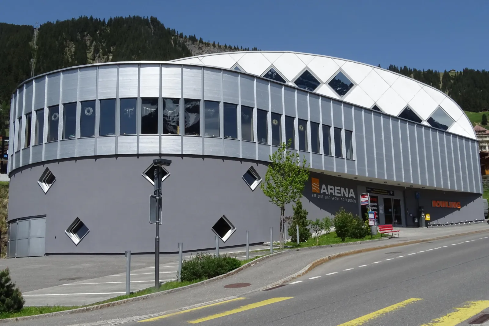 The curved facade of the Freizeit- und Sportarena Adelboden with the entrance, seen from Landstrasse — Eishalle Sportarena Adelboden
