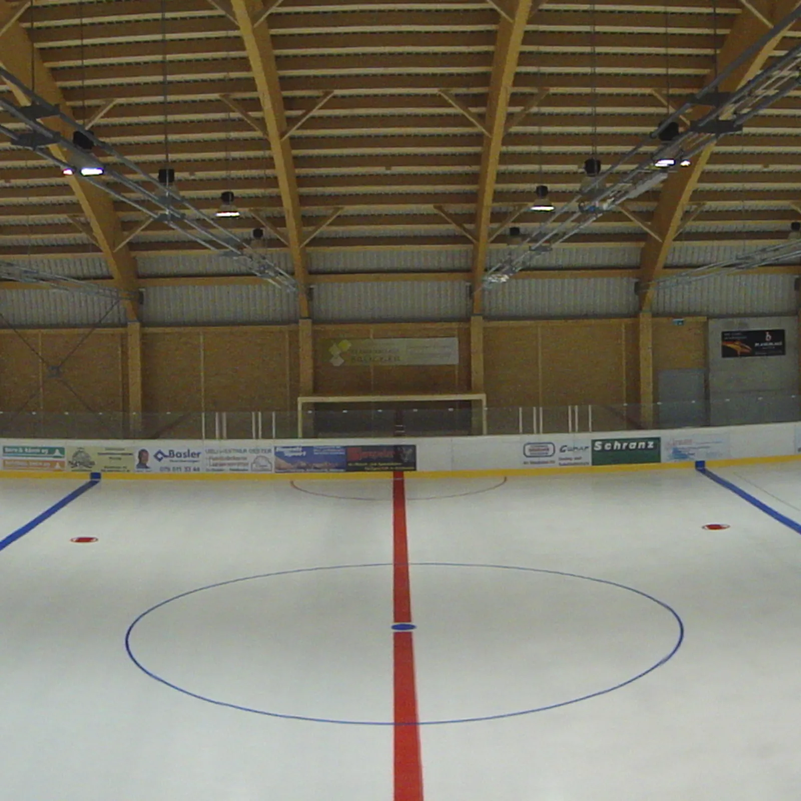 The empty ice surface of the Eishalle under the timber roof, with the centre circle and red line — Eishalle Sportarena Adelboden