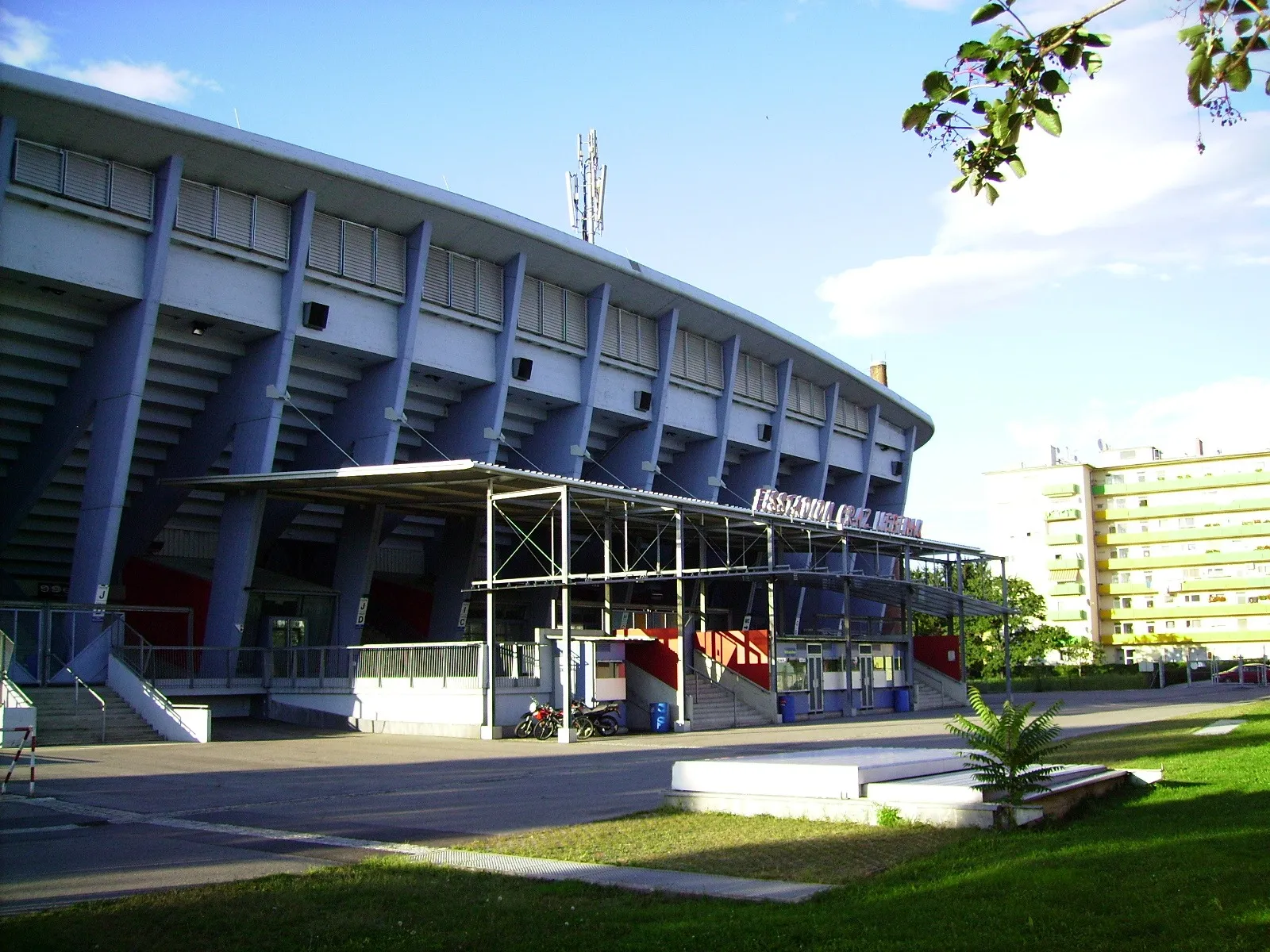 Die geschwungene blau gerippte Tribüne und die Front des Eisstadions Graz-Liebenau, darunter der überdachte Eingang und dahinter Wohnblöcke. – Eishalle B Liebenau (Merkur Eisstadion Halle B)