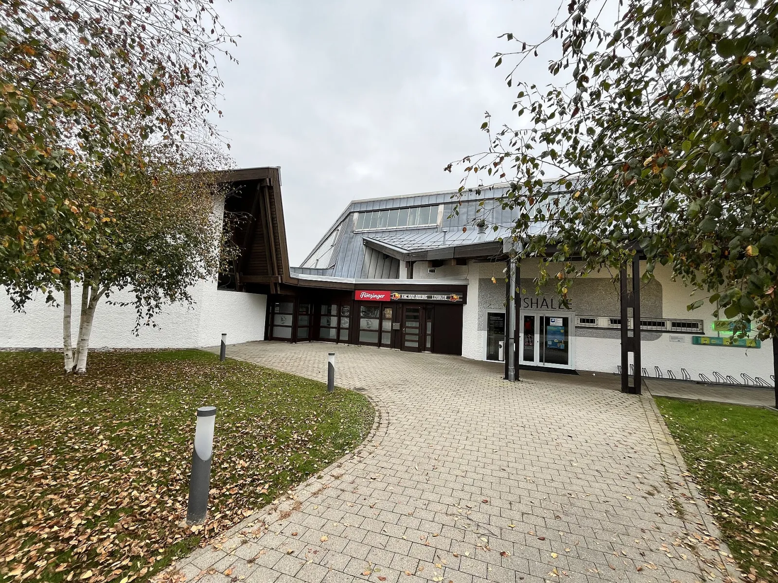 Der Eingang des Hallengebäudes vom Vorplatz aus, mit weißen Wänden, einem Baum und einem Schild mit der Aufschrift Eishalle. – Eishalle Bad Aibling