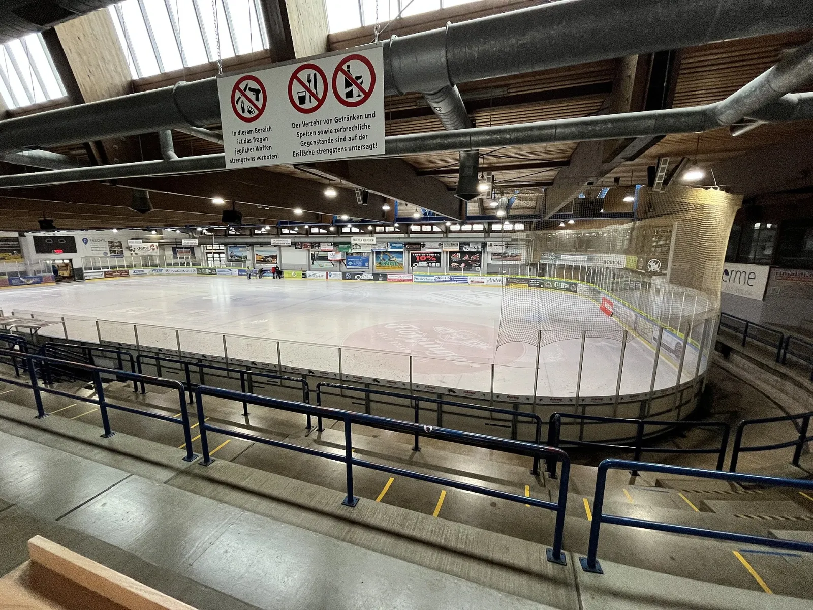 Die Eisfläche der Halle Bad Aibling von der Zuschauertribüne aus gesehen, mit Banden, Netz und leerem Eis; keine Personen. – Eishalle Bad Aibling
